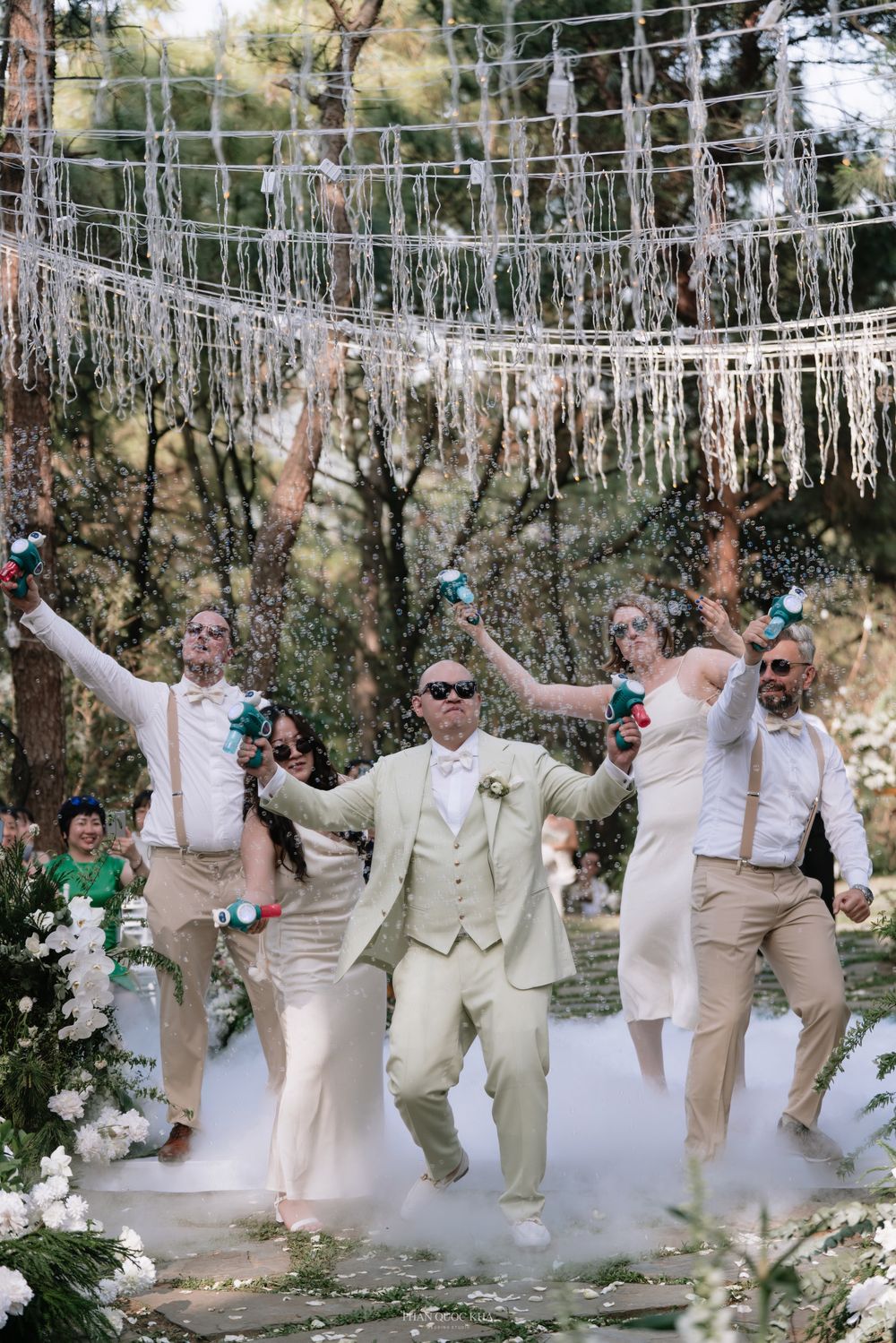 Groom and wedding party celebrate joyfully with confetti poppers under a macrame canopy at an outdoor wedding.