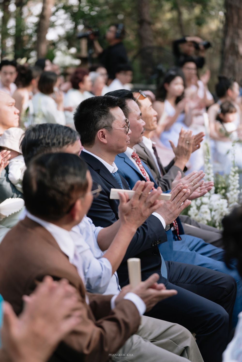 Guests clapping and applauding at an outdoor wedding ceremony seated among lush greenery.