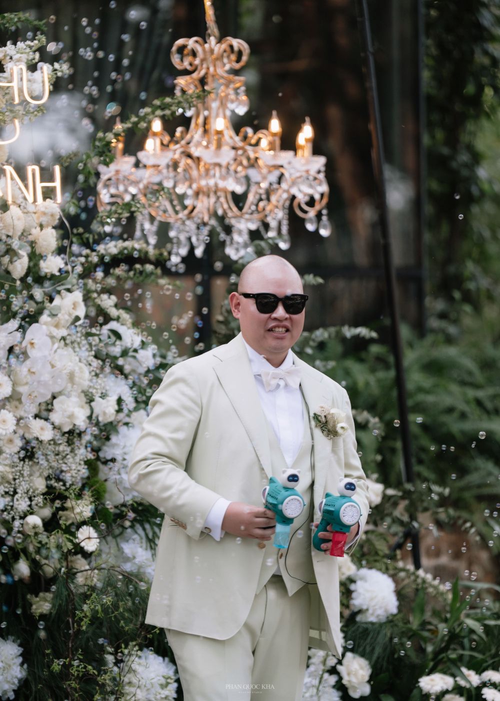 Bald man in cream suit with sunglasses holding bubble guns at a decorated outdoor wedding.