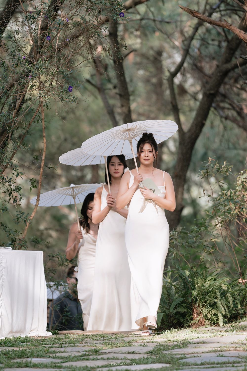 Bridesmaids in white gowns carrying parasols walking down an outdoor wedding aisle in a forest.