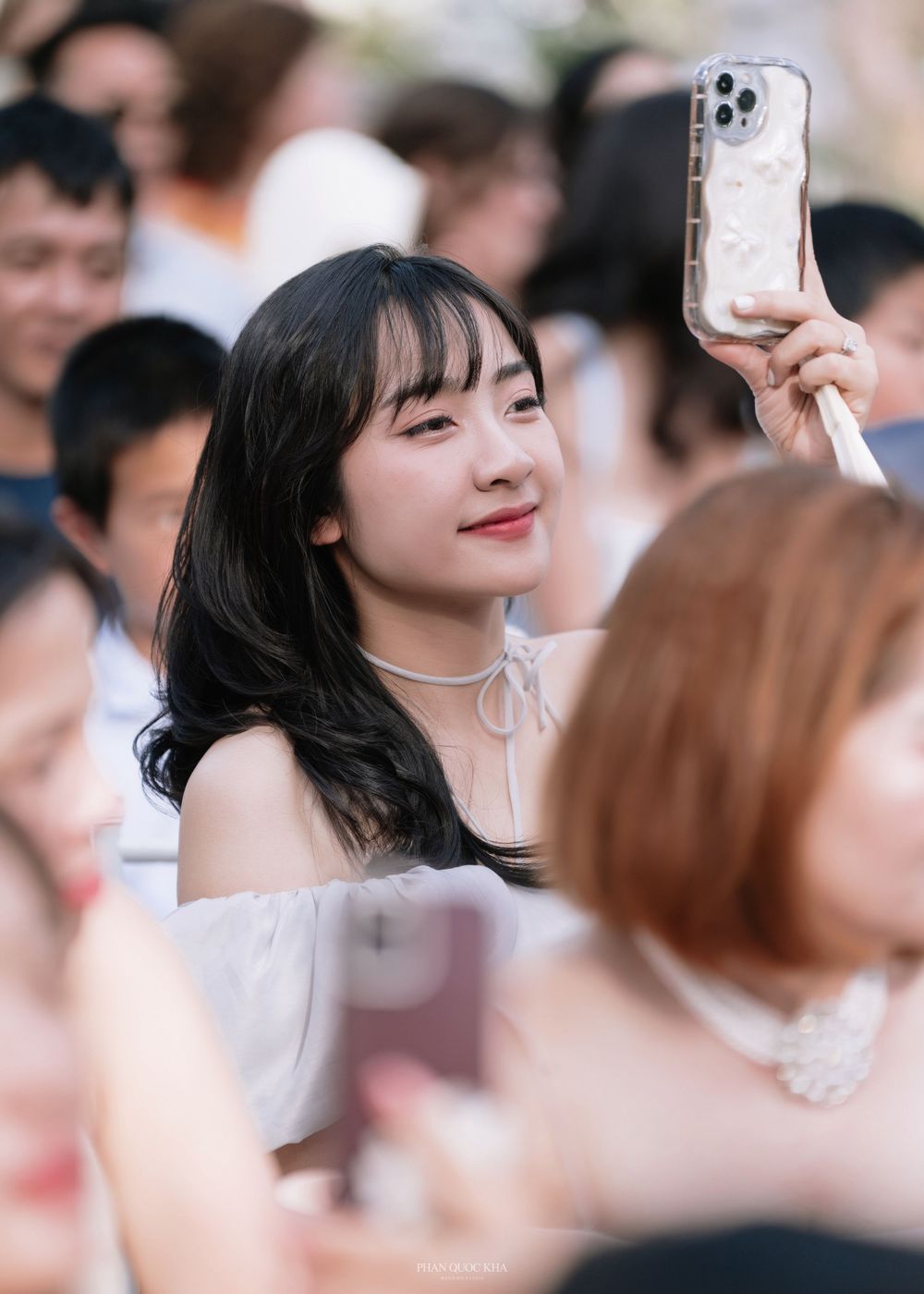 Young Asian woman smiling and taking photos on her phone at an outdoor wedding celebration.