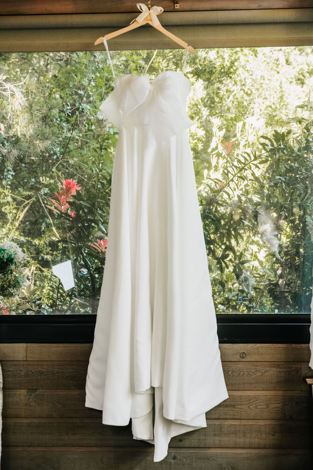 White wedding dress or gown hanging from wooden hanger against rustic wood panel wall with green foliage visible outside.