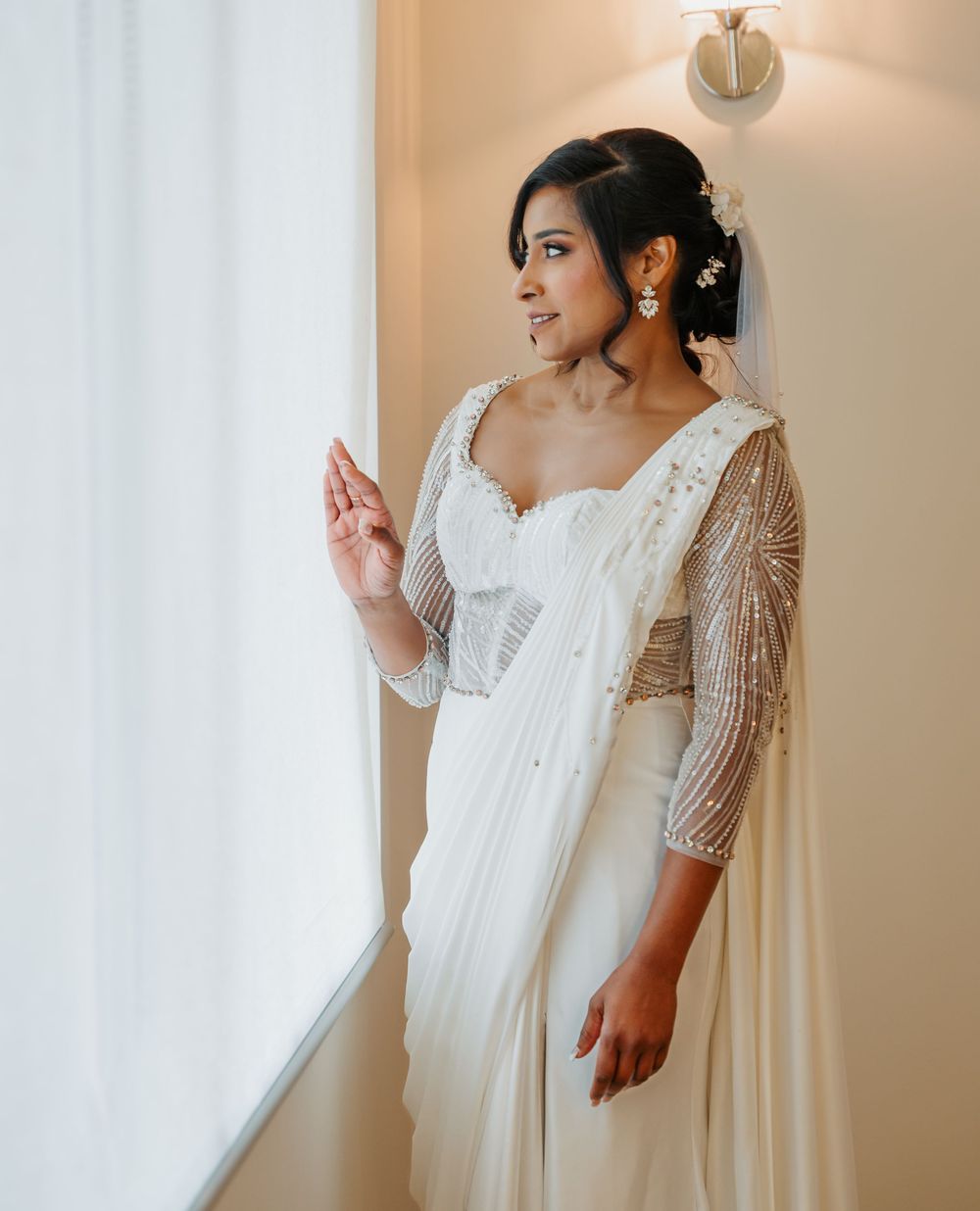 A person in a white flowing wedding dress with beaded sleeves poses elegantly against a light backdrop.