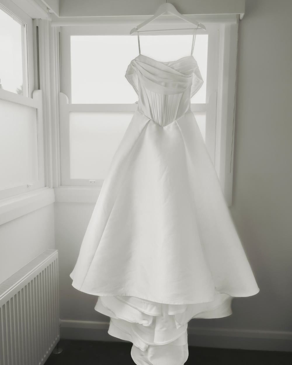 A white wedding dress with a fitted bodice and full skirt hangs gracefully near a window in soft natural light.
