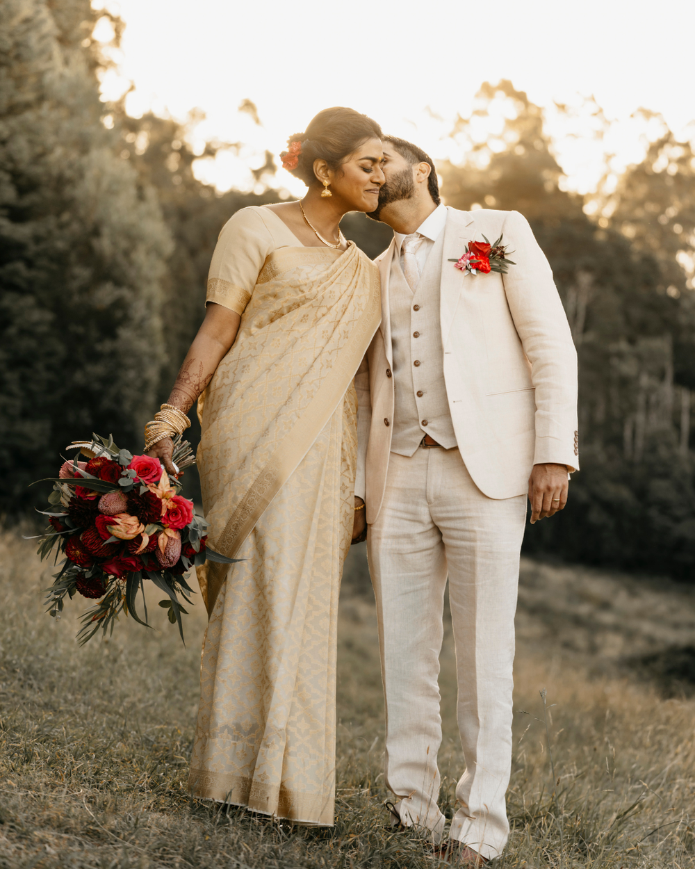 A couple in beige attire poses intimately in a golden meadow holding red flowers at sunset.