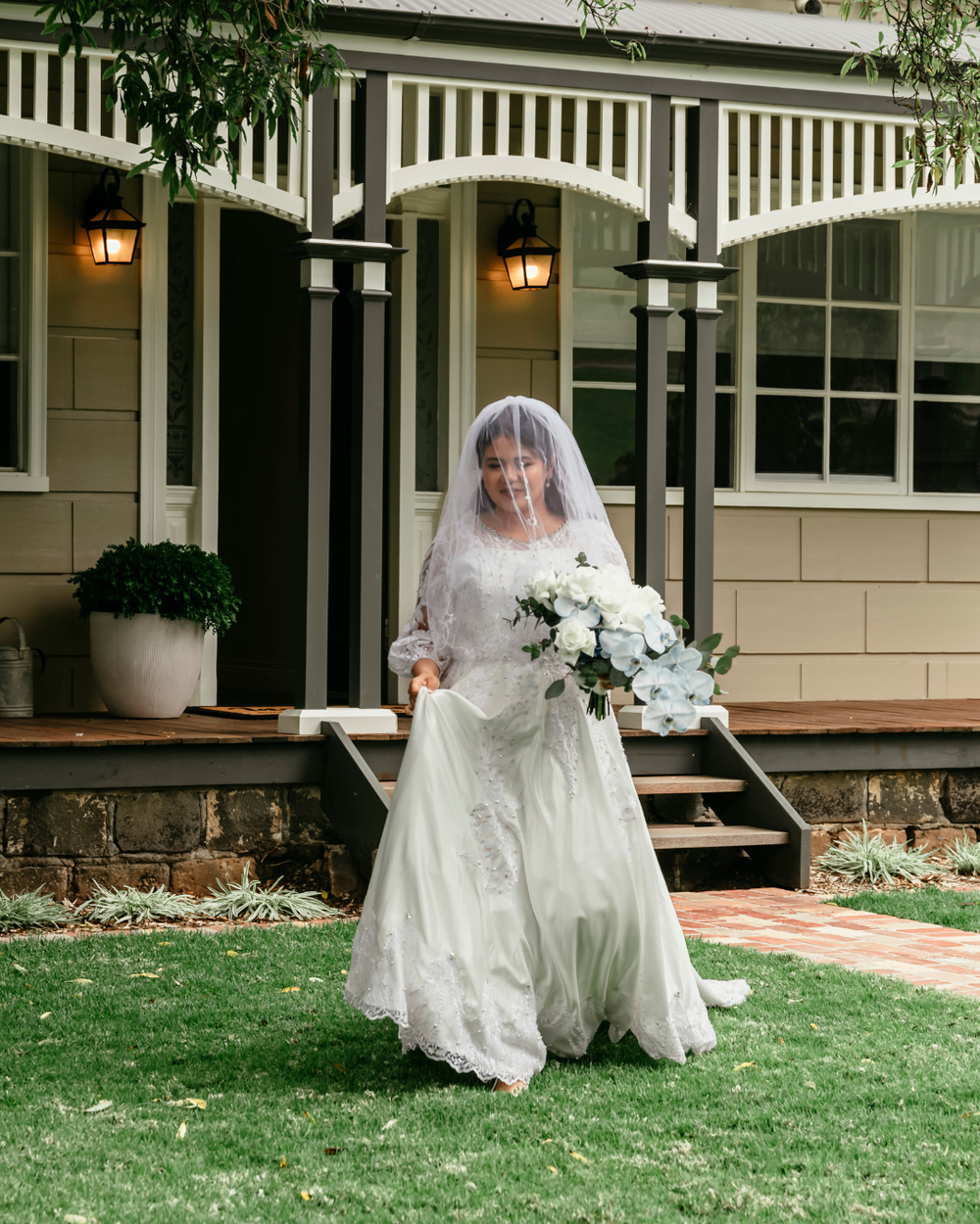 A bride in a white lace wedding gown sits gracefully on the lawn outside a Victorian-style house.