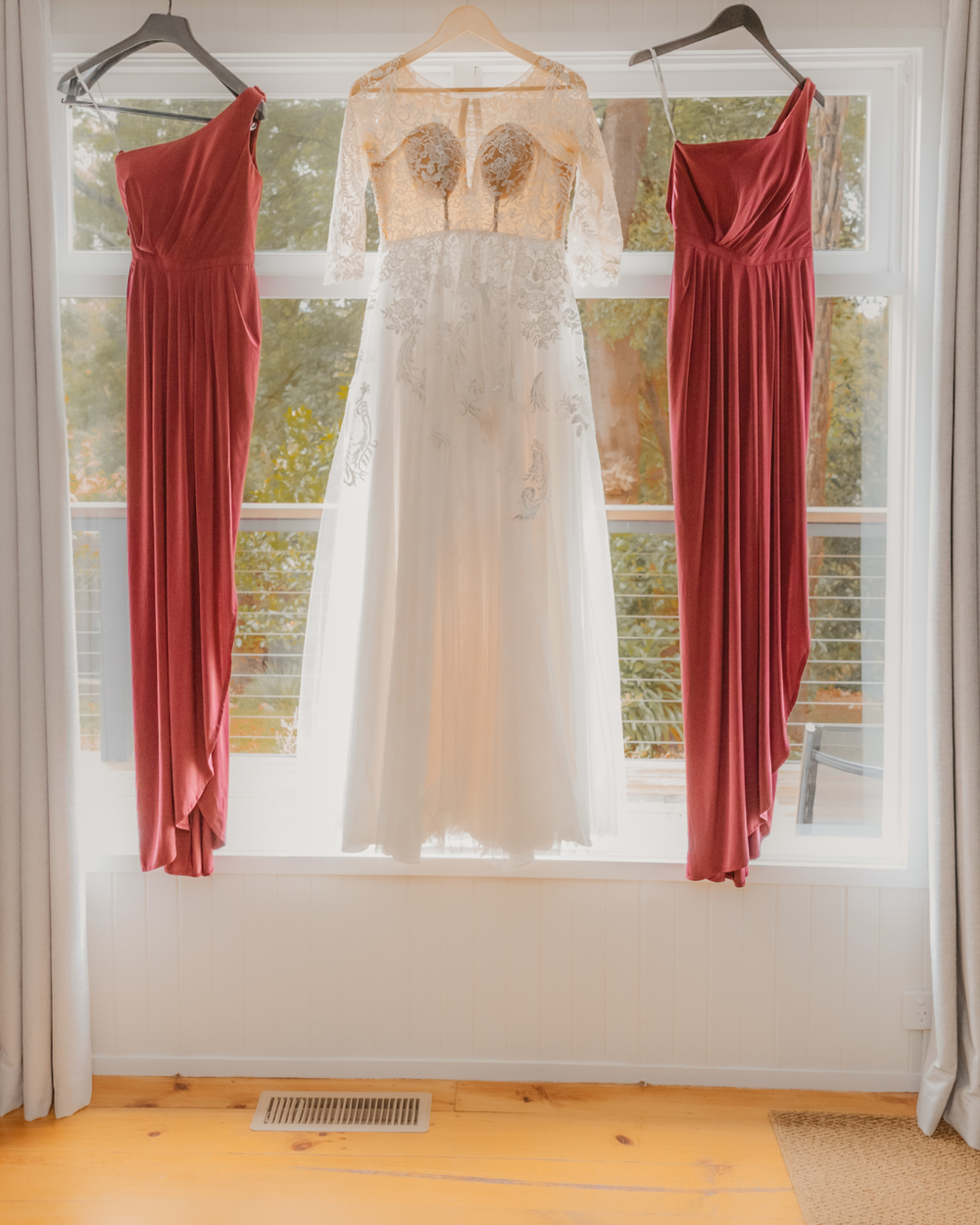 Elegant wedding dresses and burgundy bridesmaid gowns hanging on display in a sunlit room with white curtains.