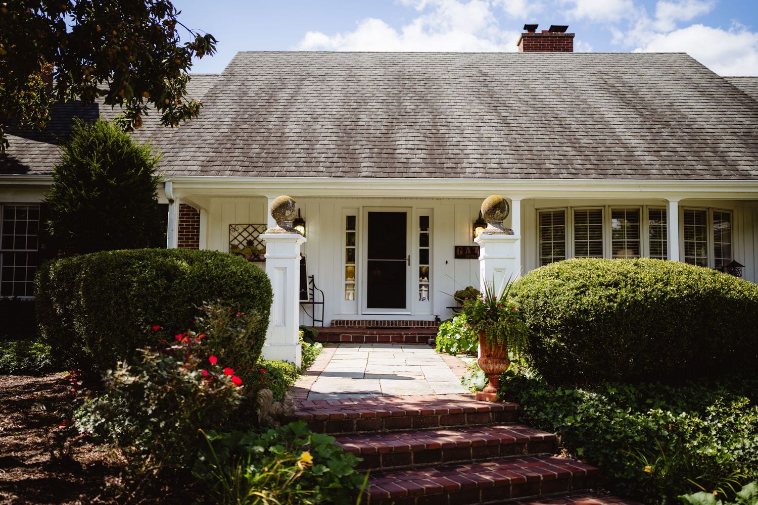 Two-story traditional brick house with dormers and white trim against a blue sky and manicured landscaping.