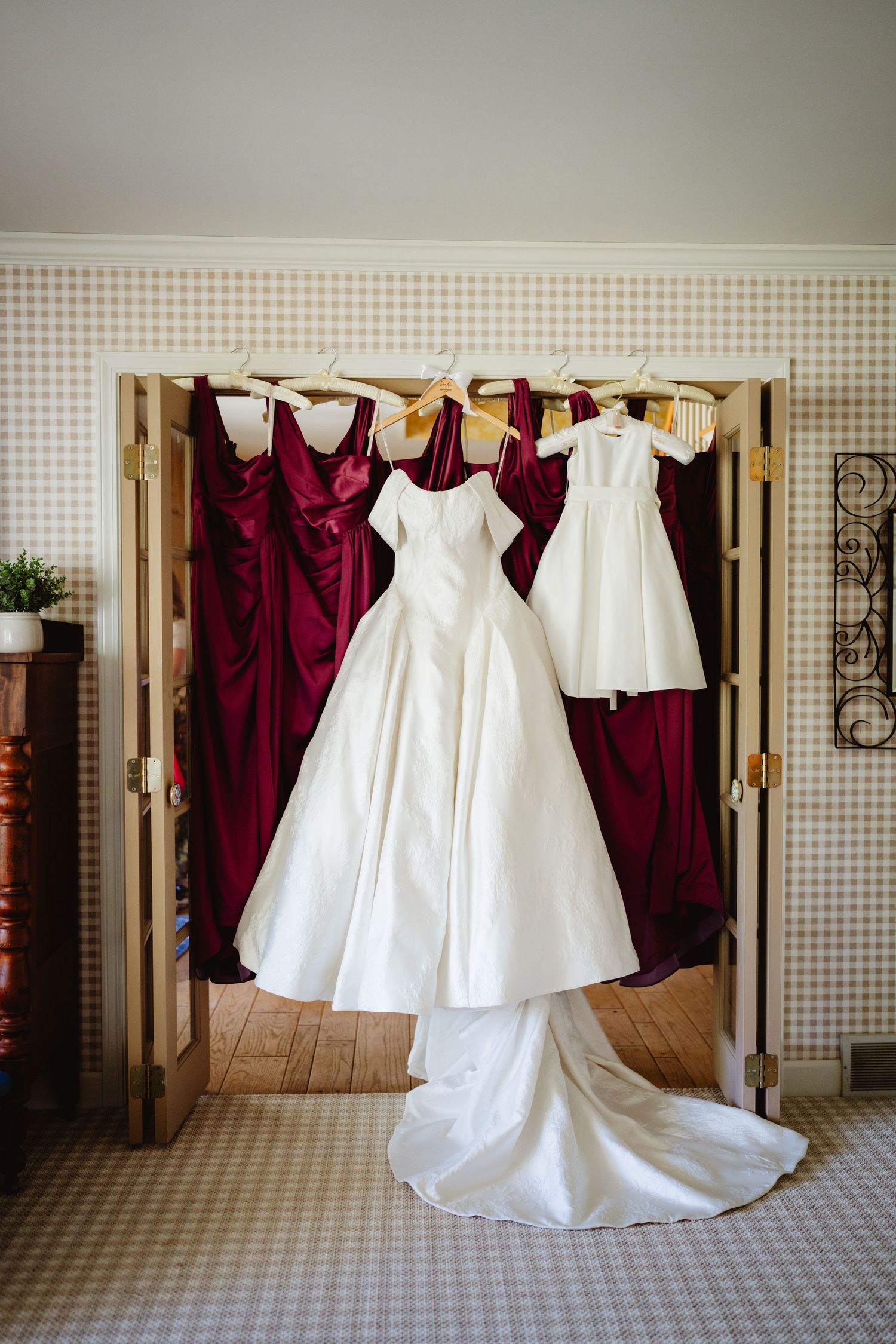 White wedding gown and burgundy bridesmaid dresses hanging on a wooden folding door in a decorated room.