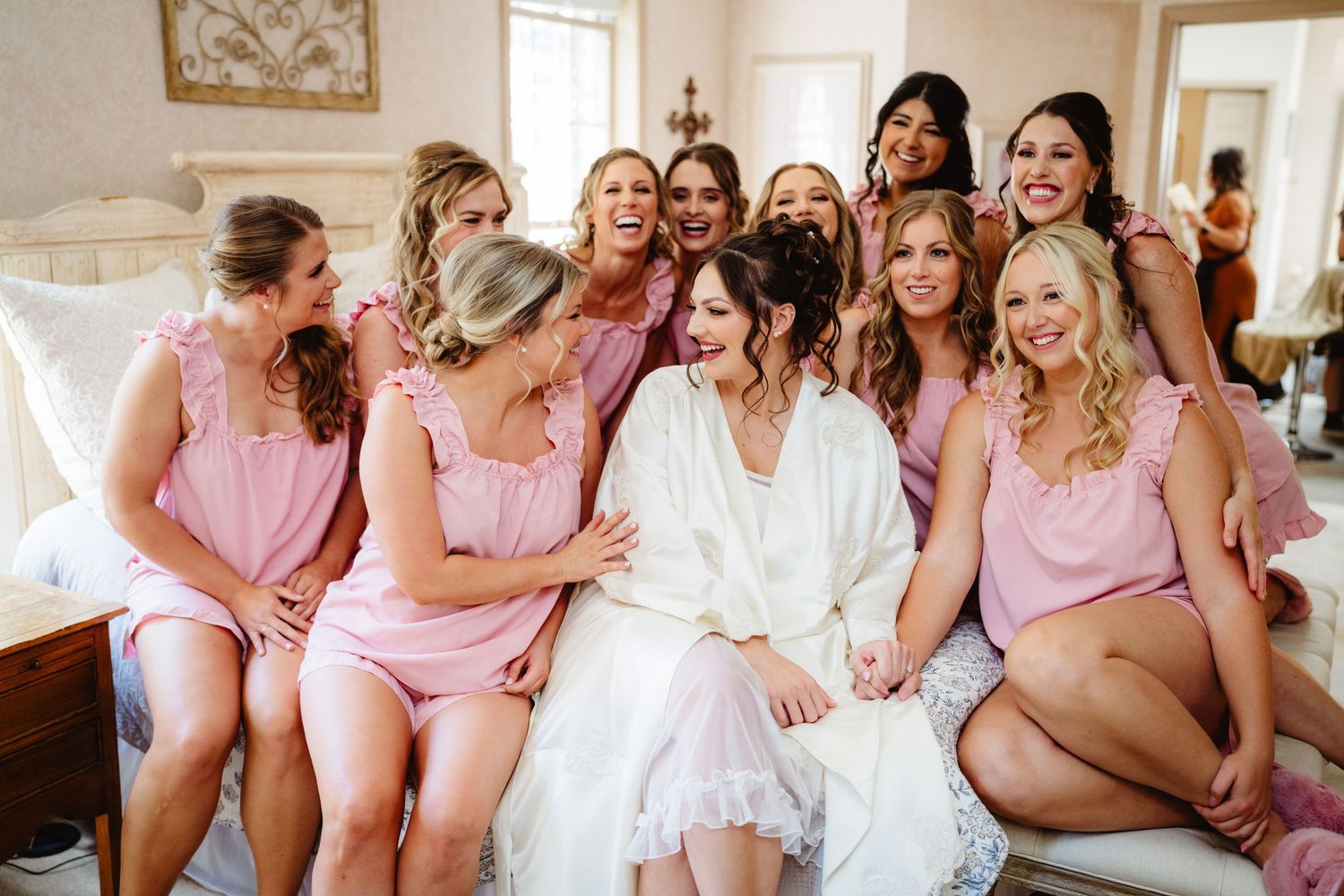 Group of bridesmaids in matching pink robes laughing together on wedding day morning.