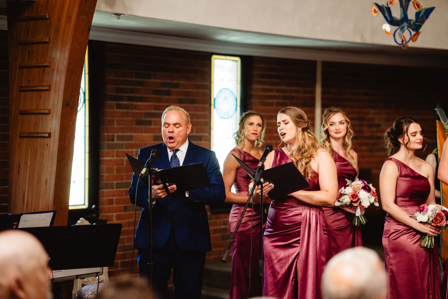Wedding party in burgundy bridesmaid dresses and black tuxedos laughing together outside a brick building.