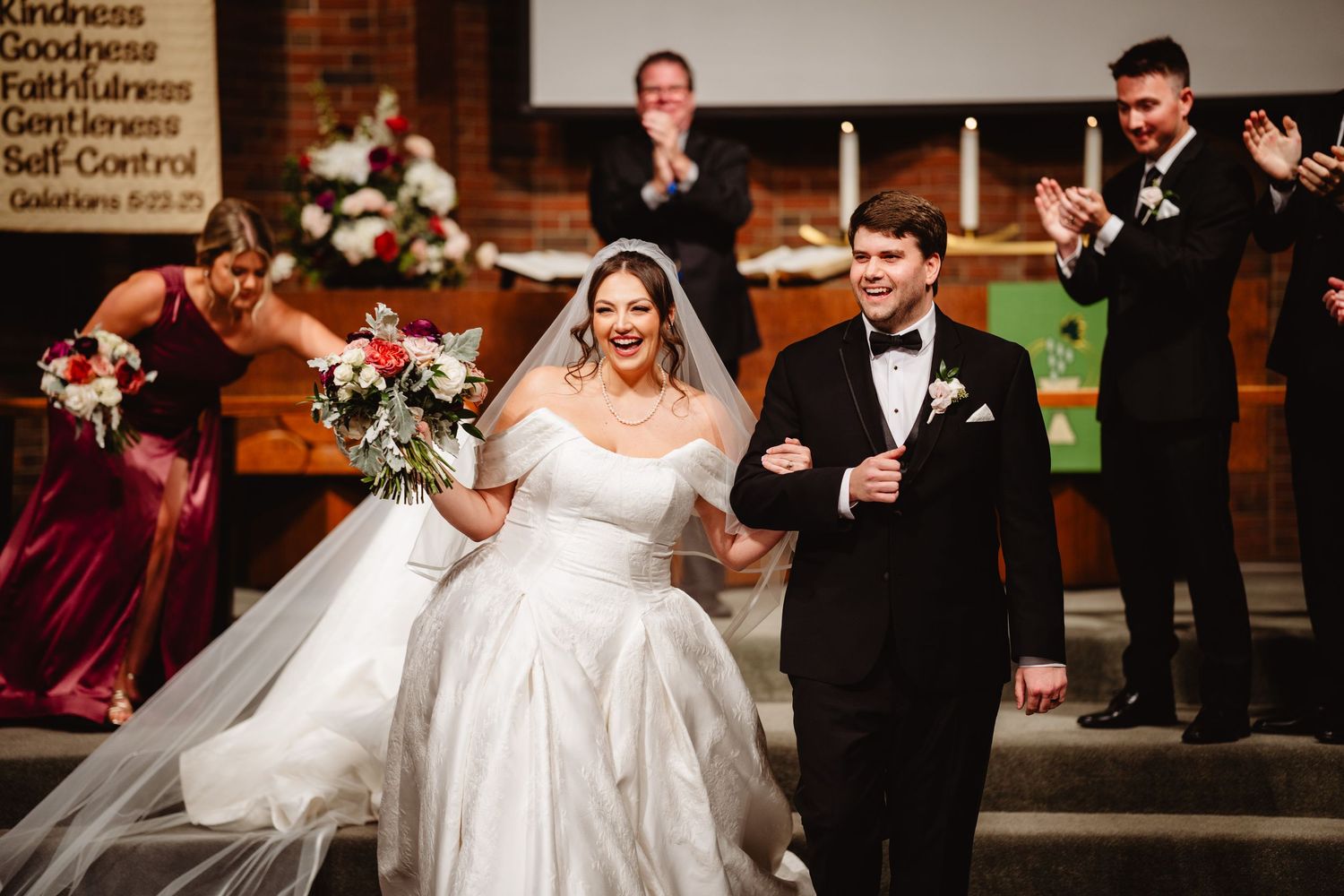 Wedding ceremony with bride in white gown and groom in black tuxedo at a brick-walled church.