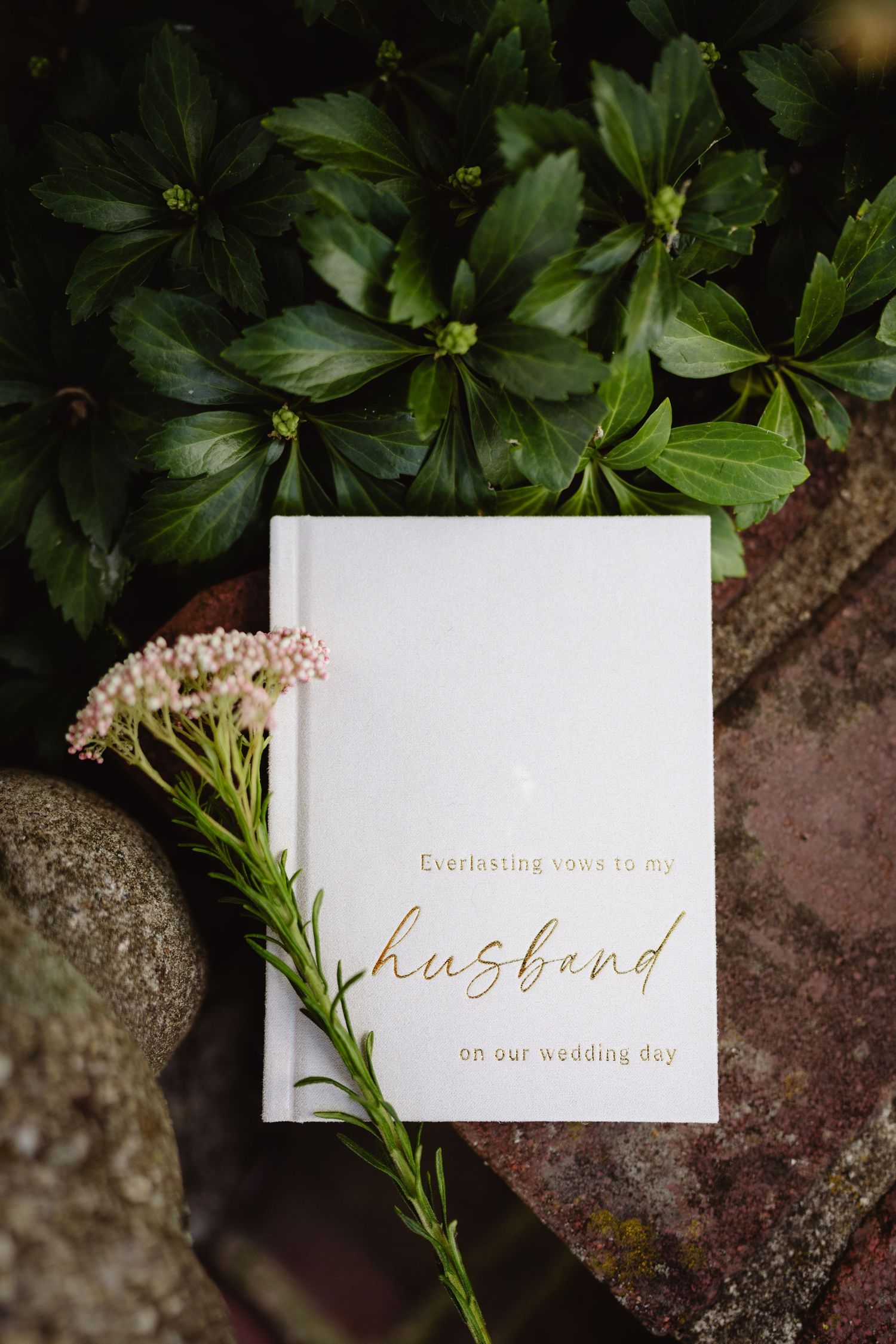 White wedding card with gold text and green foliage decorations against a rustic stone background.