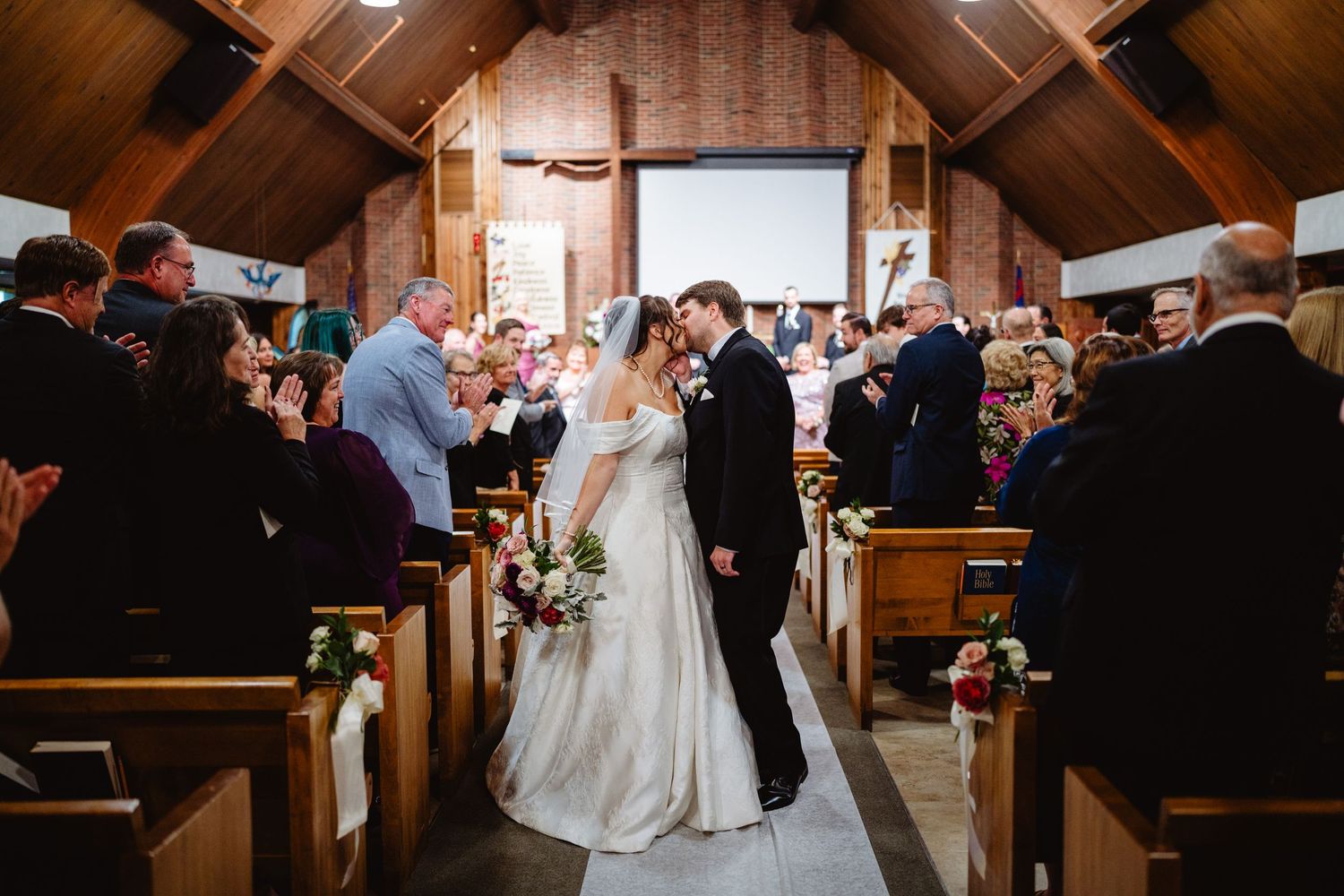 A wedding ceremony takes place in a rustic wooden chapel with guests seated in pews as natural light streams through windows.