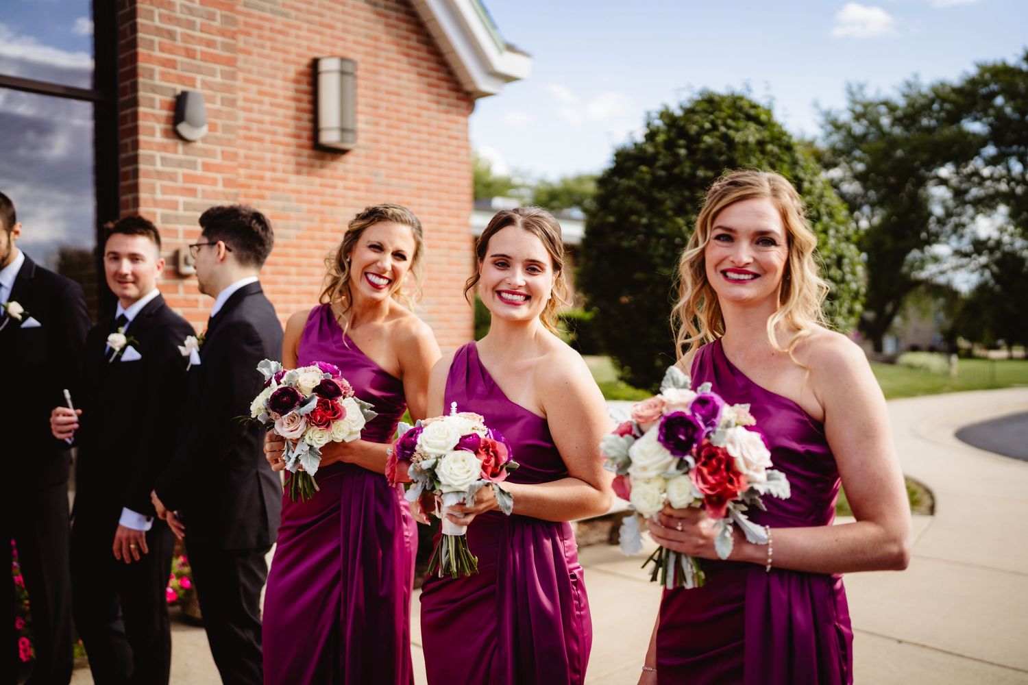 Bridal party wearing burgundy dresses posing for photos in front of brick church on sunny day.