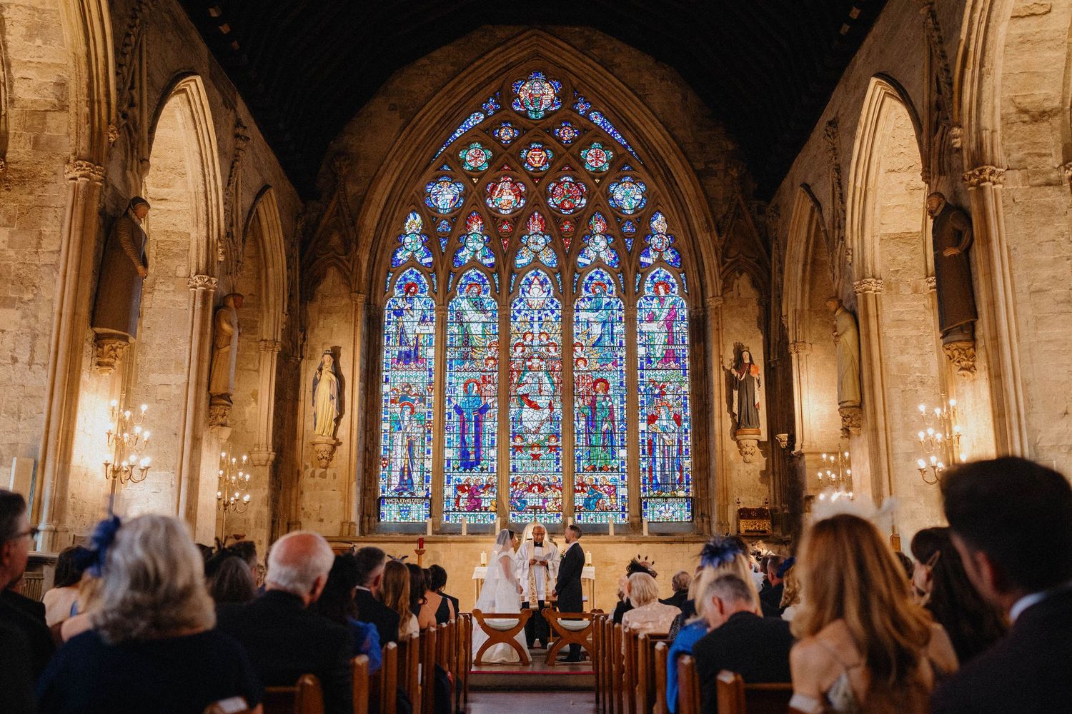 Wedding ceremony in St Etheldreda's church with stained glass window, stone arches as seated guests in wooden pews watch the couple at altar