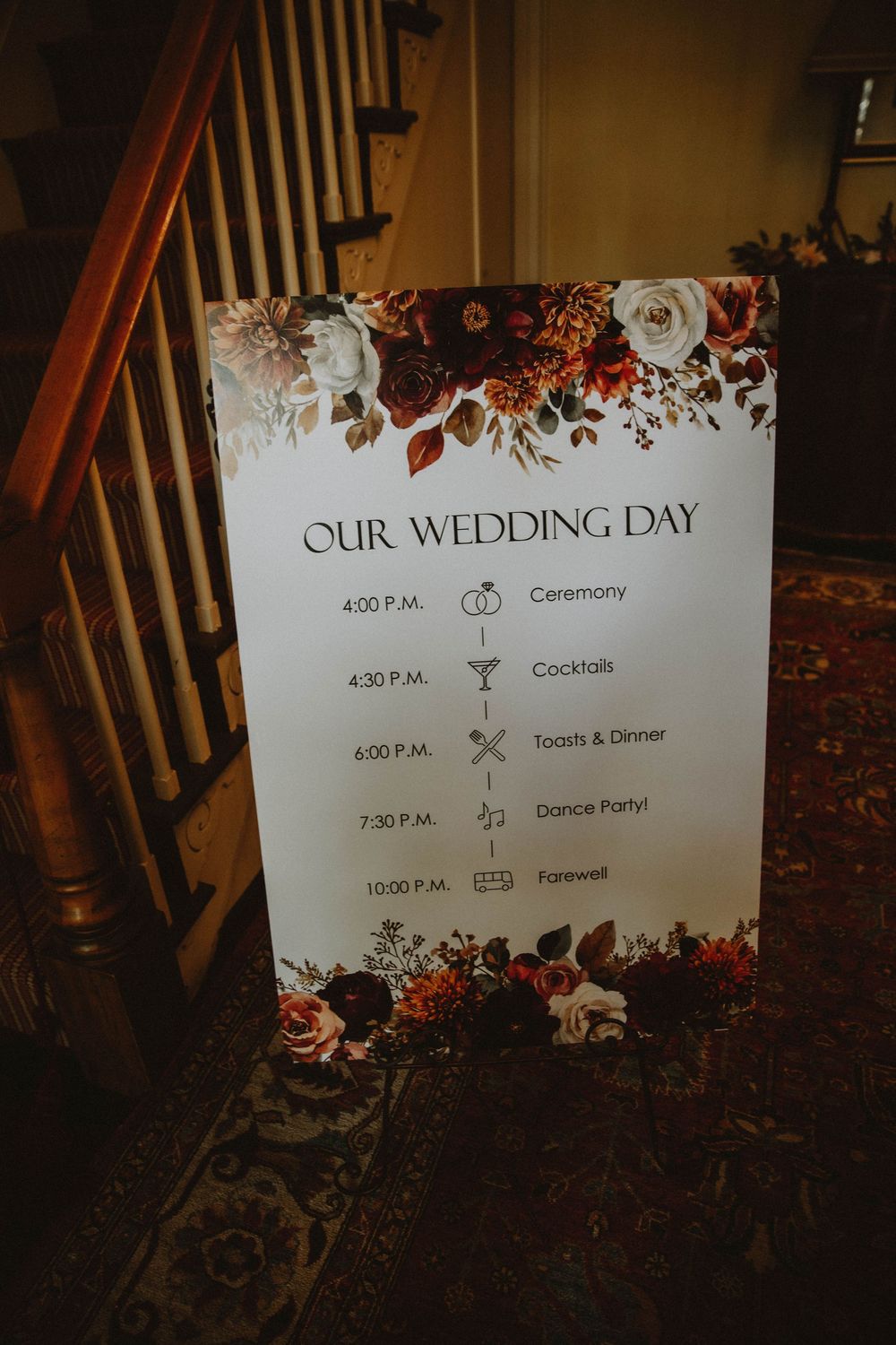 Rustic wedding timeline sign for ceremony and reception displayed over autumn floral decorations and dark wood floor.