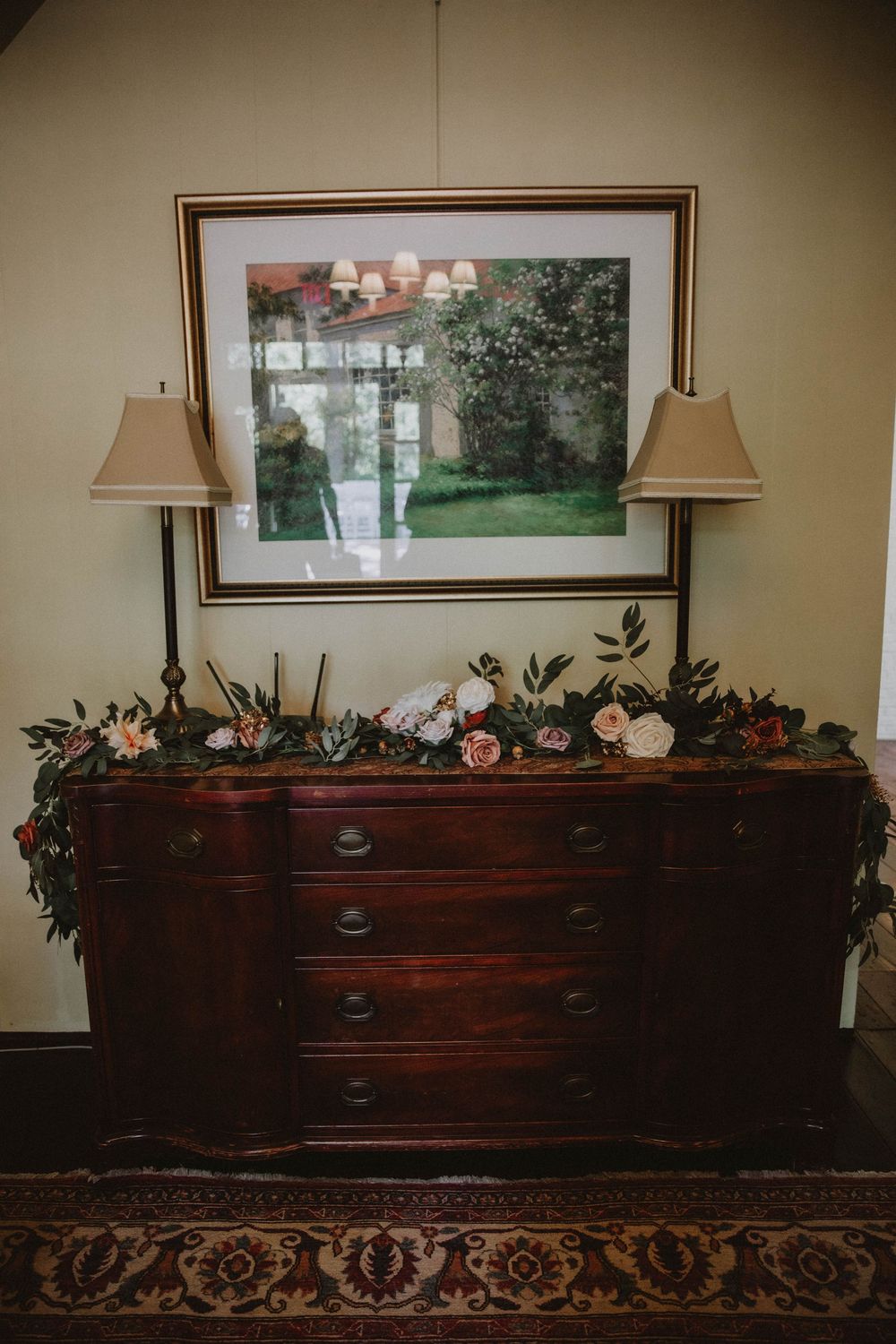 Vintage wooden chest decorated with ornate carvings and greenery displays sits against beige wall under framed artwork.