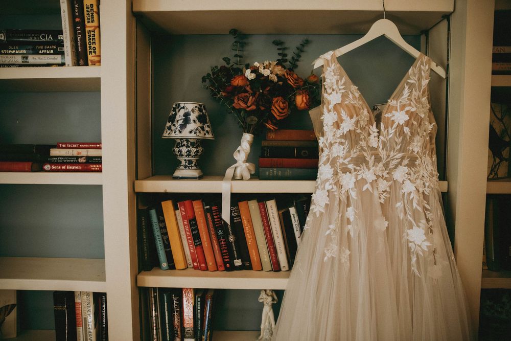 Delicate lace wedding dresses displayed on hangers against bookshelves in a bridal boutique setting.