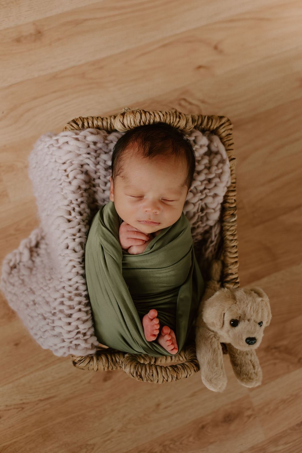 Sleeping newborn swaddled in green fabric with stuffed puppy toy on wooden floor.