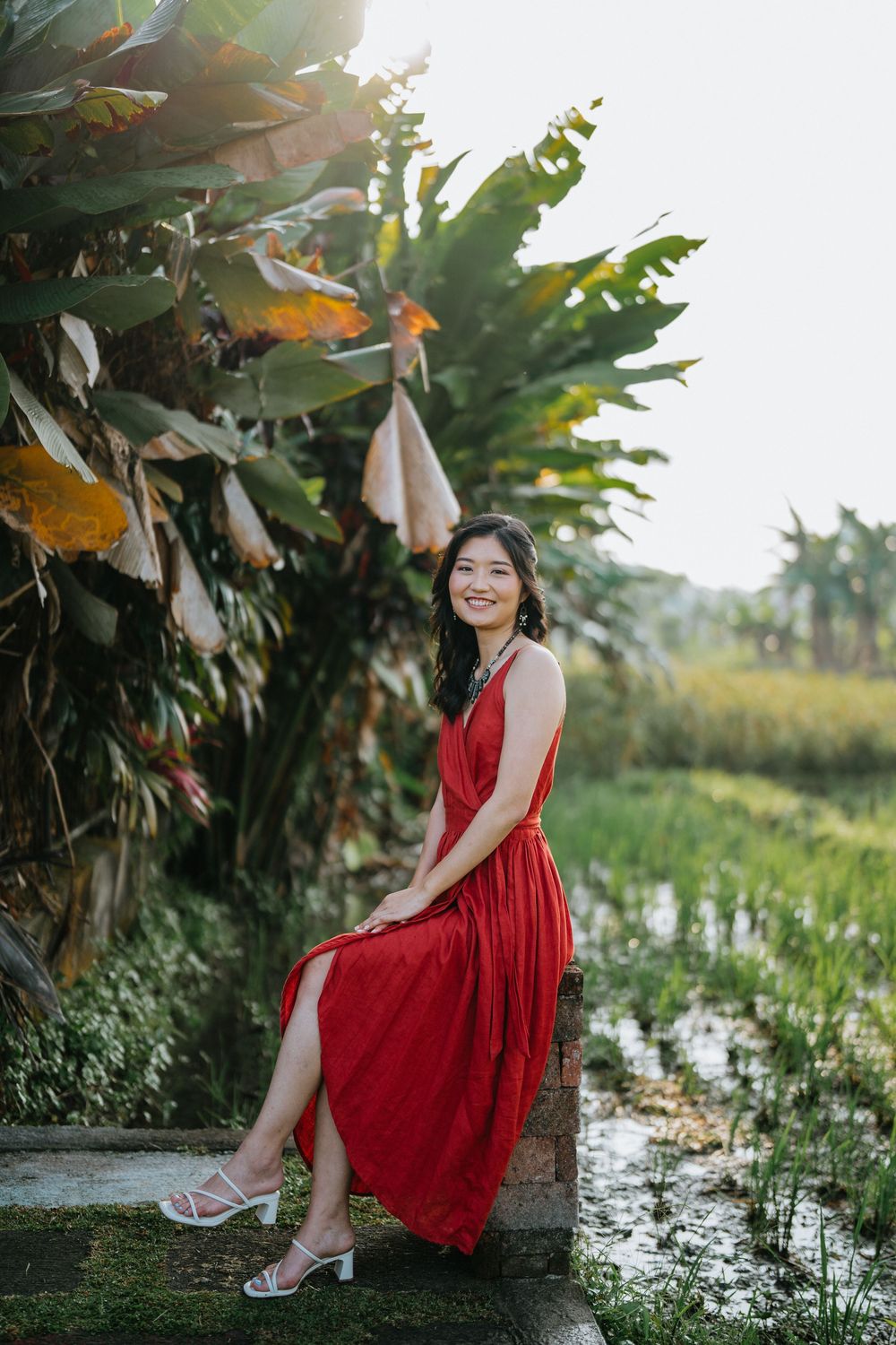 Woman in flowing red dress poses elegantly in lush tropical garden setting.