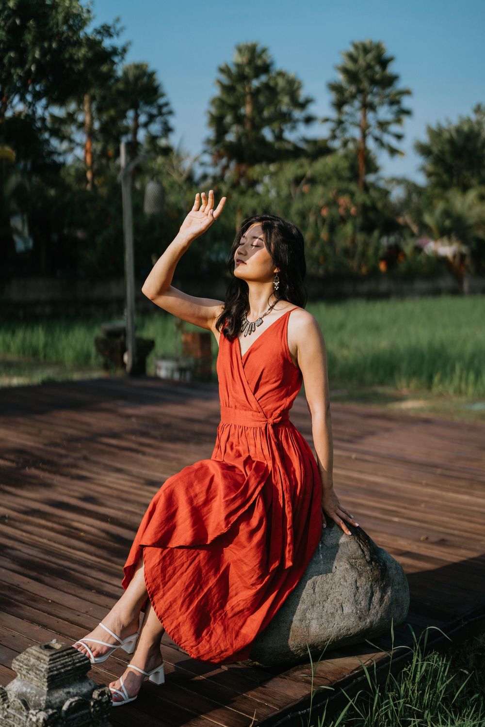 Woman in red dress sits gracefully on rock amid tropical garden landscape.