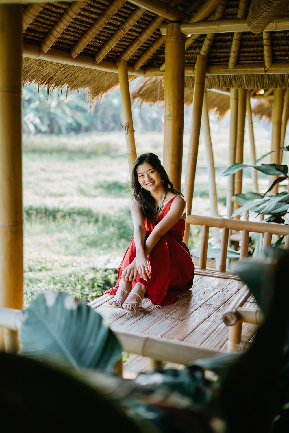A red dress contrasts beautifully against a rustic bamboo hut setting with thatched roof and natural lighting.