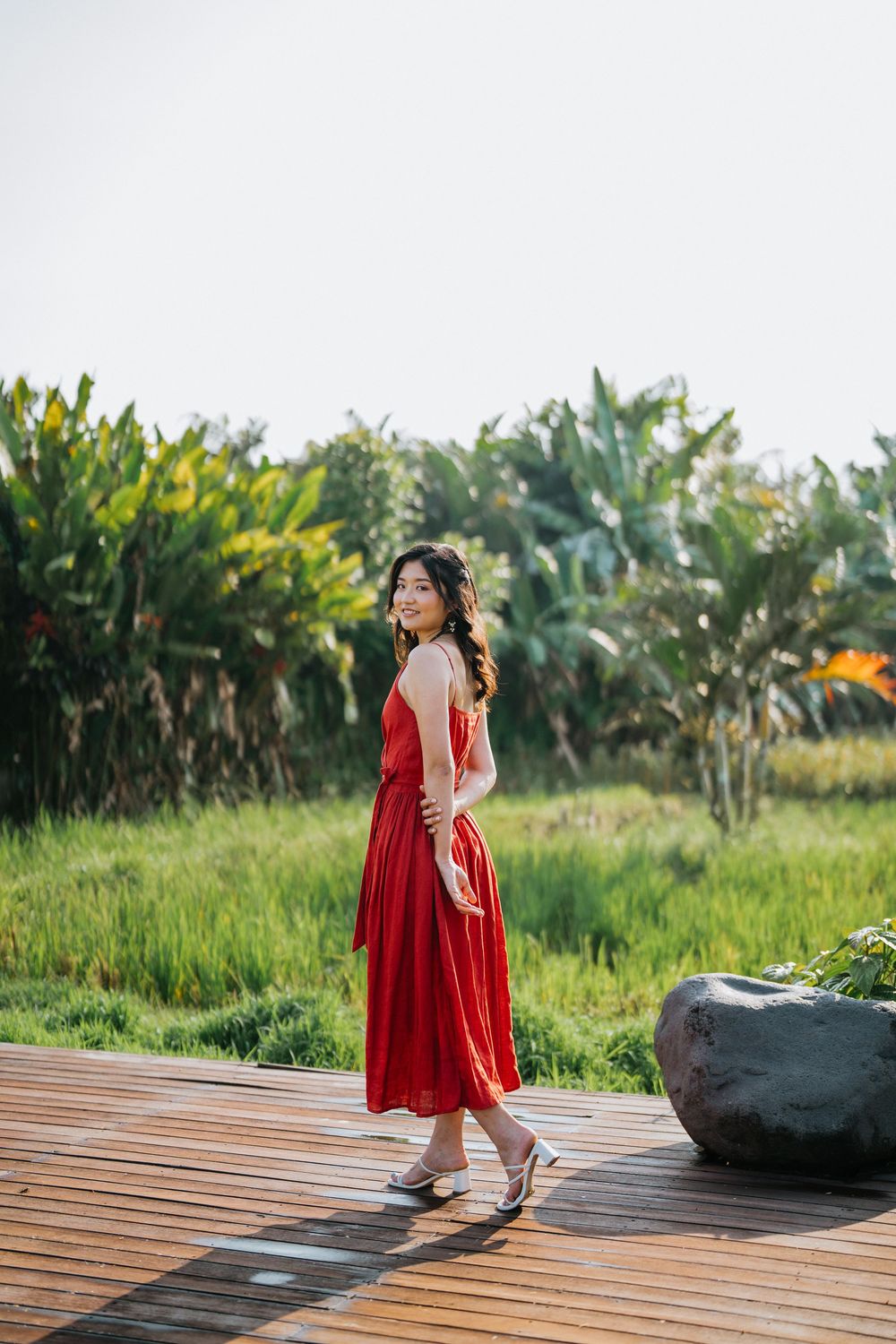 A red sundress stands out on a wooden deck pathway surrounded by lush tropical gardens.