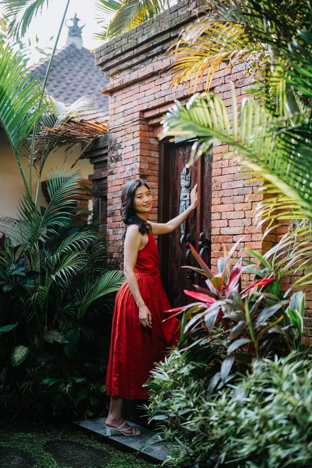 Red dress adds vibrant color against rustic brick doorway surrounded by tropical plants.