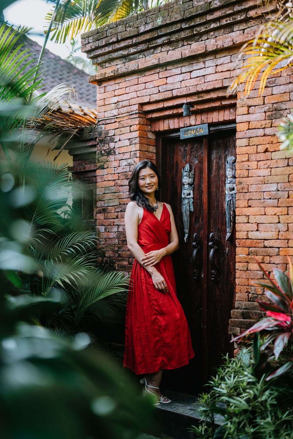 Elegant pose by brick doorway with red dress complementing the tropical garden surroundings.