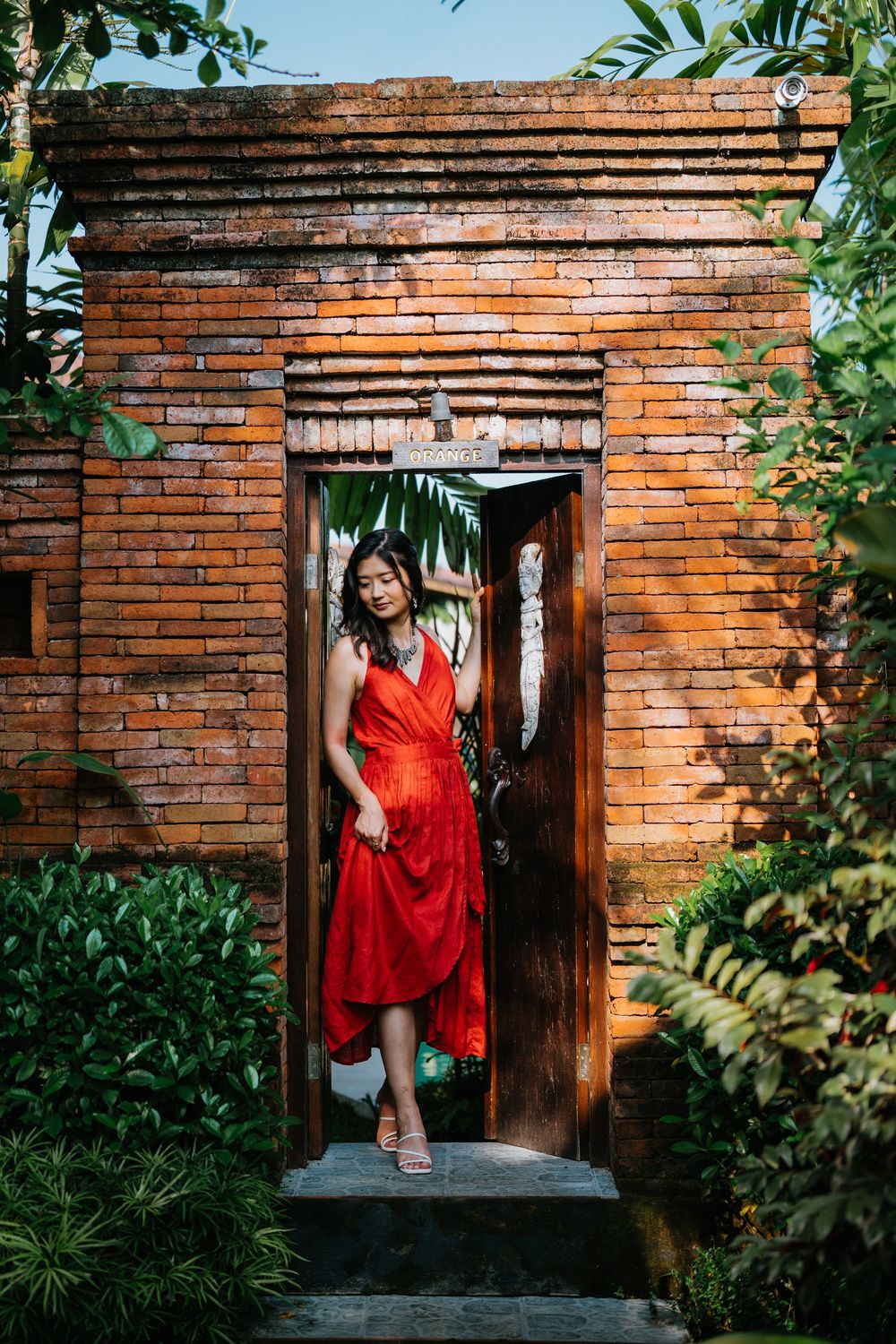 Red dress creates striking contrast against weathered brick entrance and lush garden foliage.