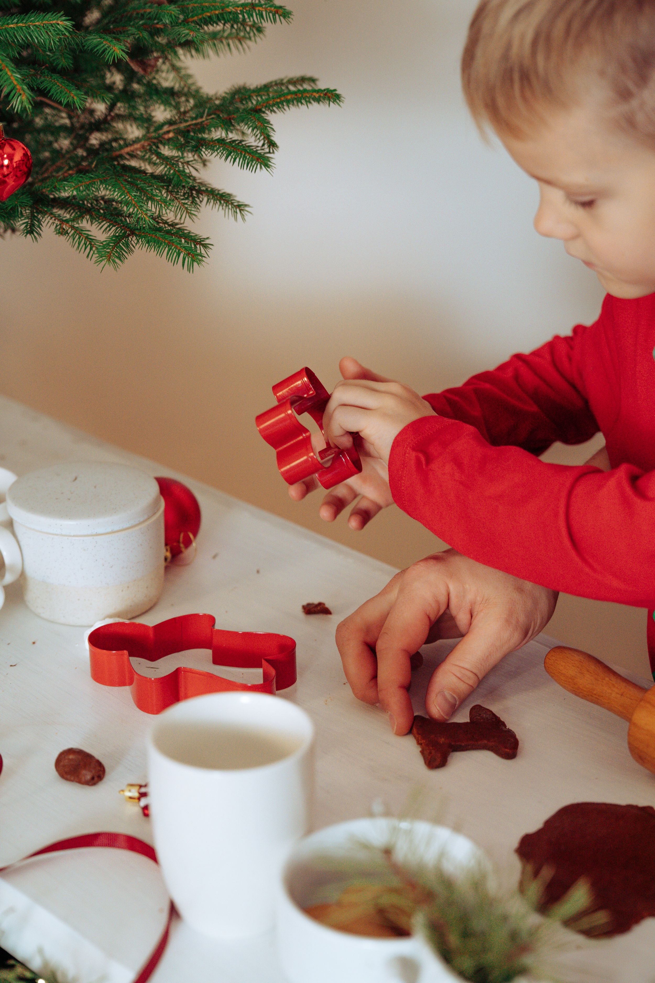 Close-up of hands using red cookie cutters to make Christmas cookies on a white surface.