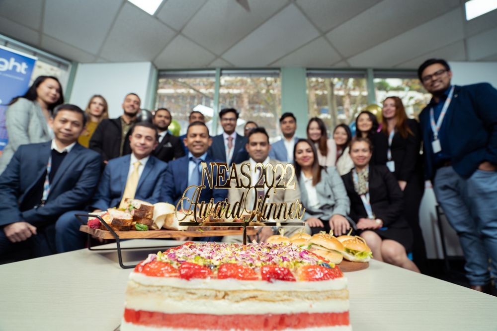 A group gathers around a decorated birthday cake with 'world line' text topper in an office meeting room.