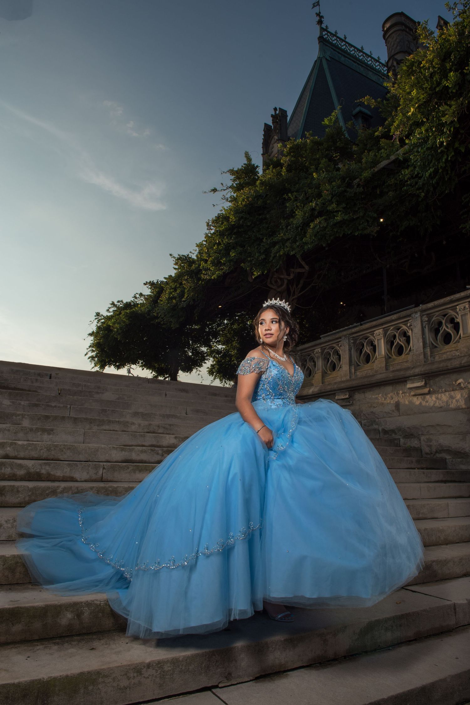 A person in a flowing light blue ball gown poses on stone steps at dusk.