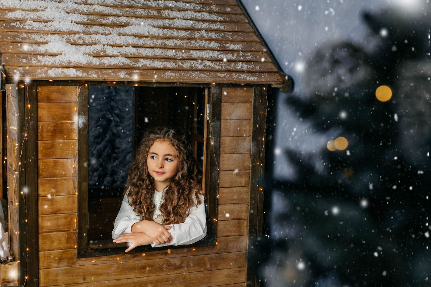 Retrato de una niña disfrutando de una sesión de fotos navideña mirando por la ventana de la casita de navidad.