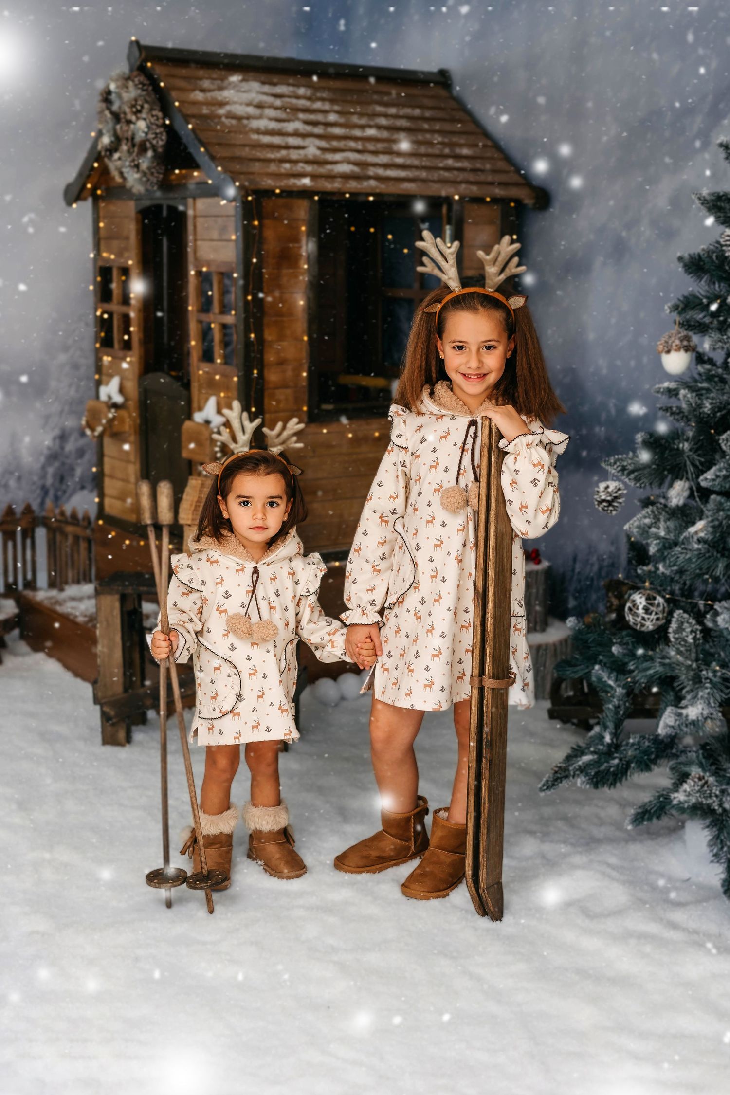Retrato de dos niñas disfrutando de una sesión de fotos navideña con los esquís y casita navideña de fondo.