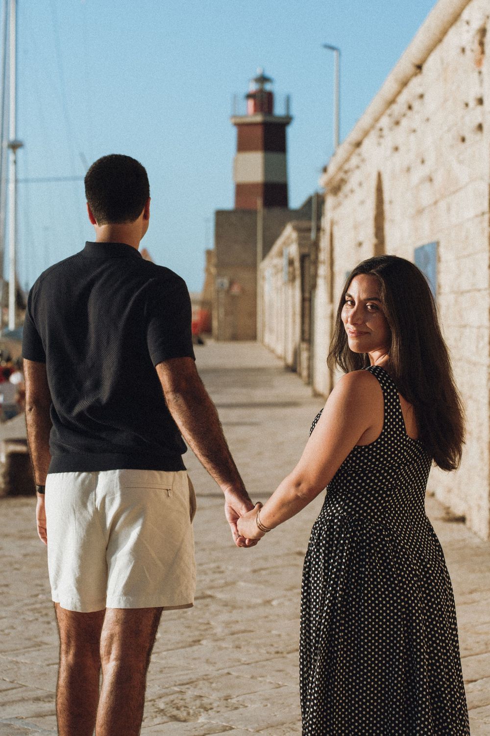 A couple strolls along a sunny stone walkway with a lighthouse visible in the distance.