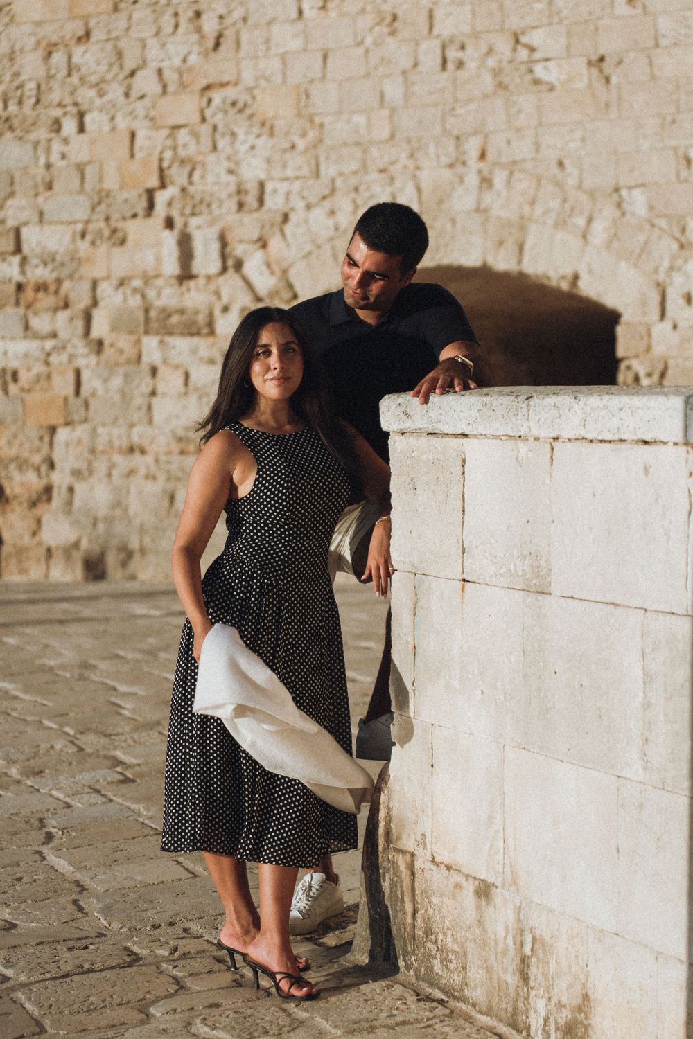 Woman in polka dot dress sits on stone steps against ancient beige wall in Mediterranean setting.