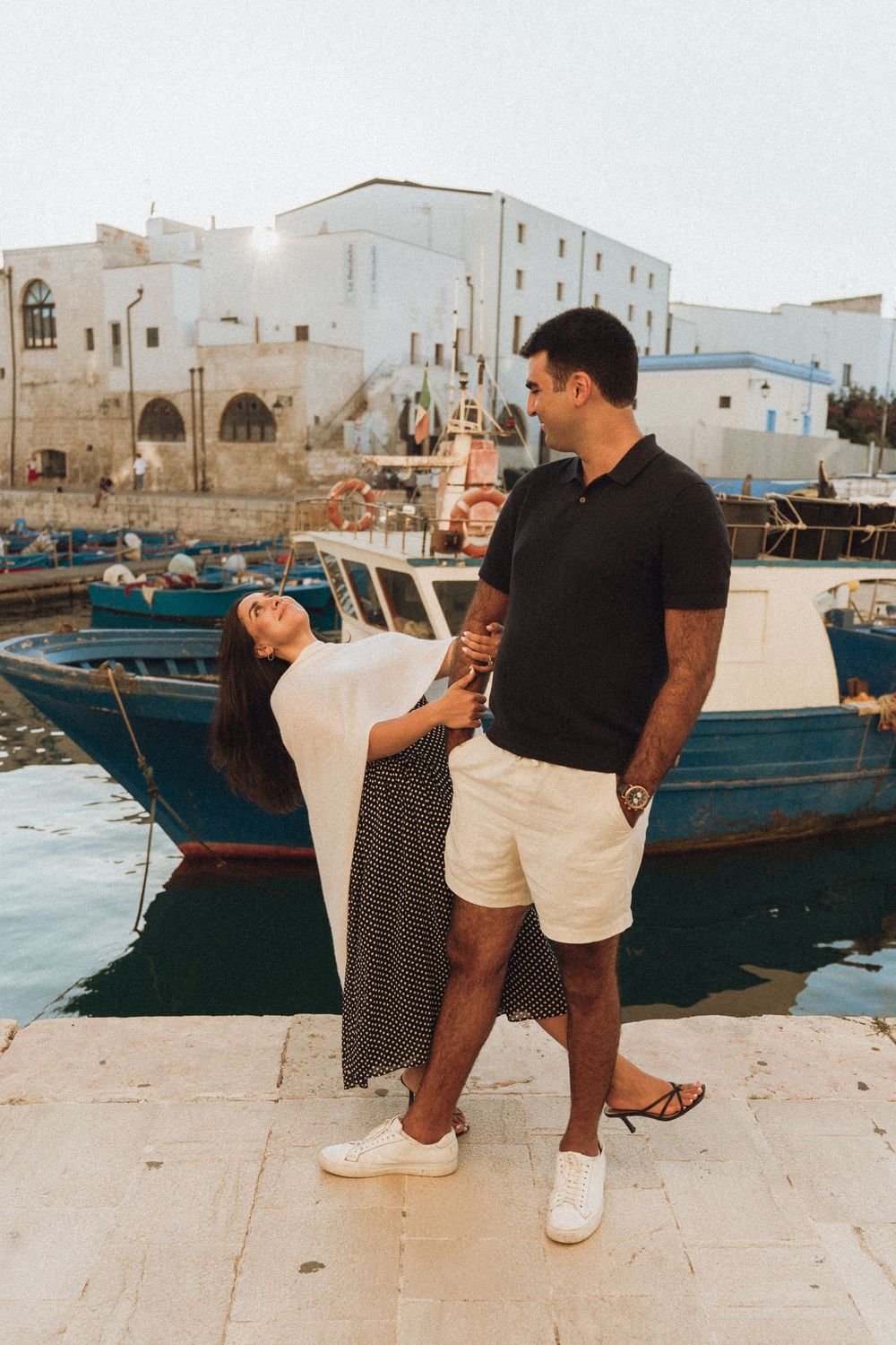 A romantic couple takes playful photos by the marina with wooden boats and white buildings in the background.