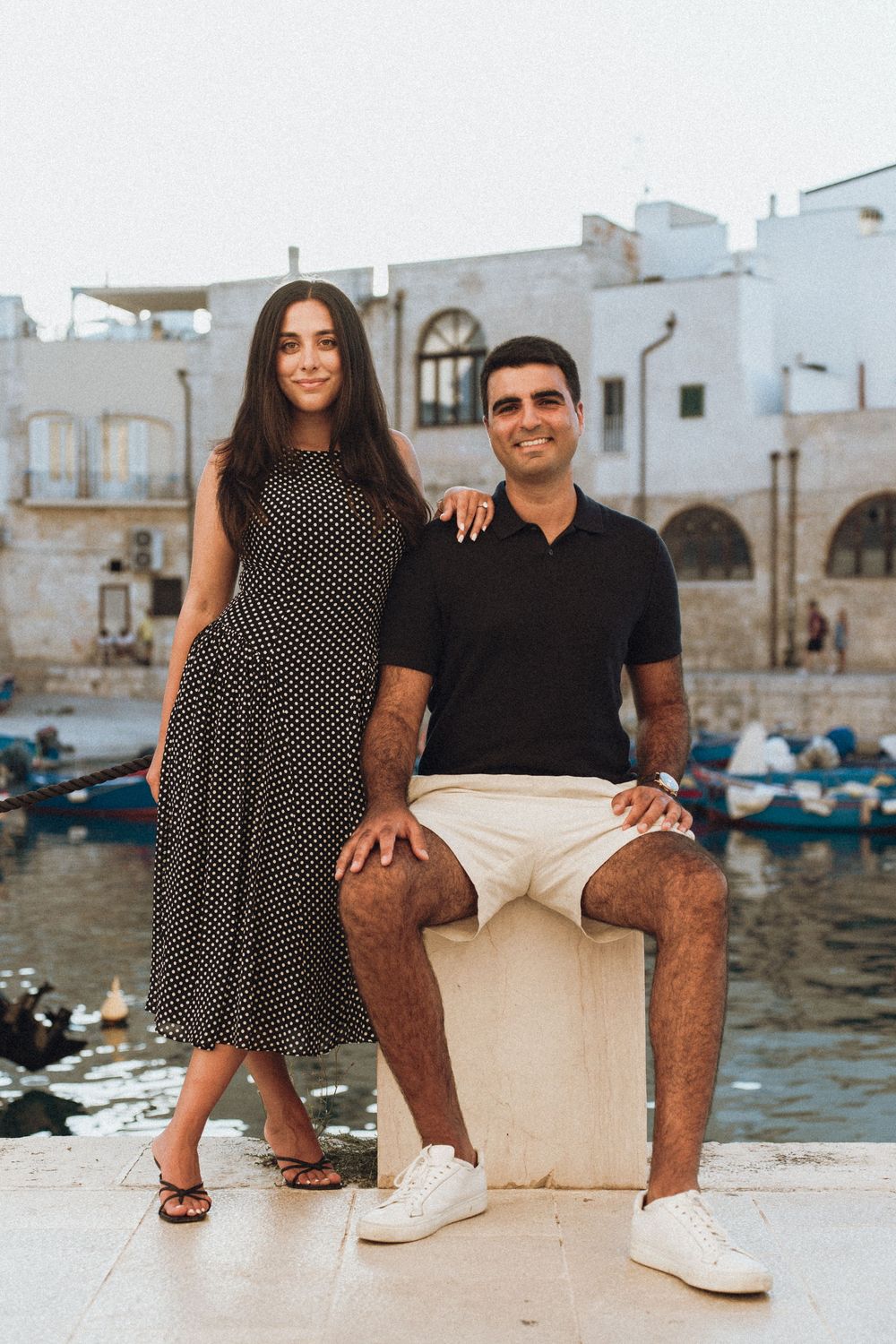 A couple poses along the waterfront with boats and white buildings visible in the background near the Mediterranean sea.