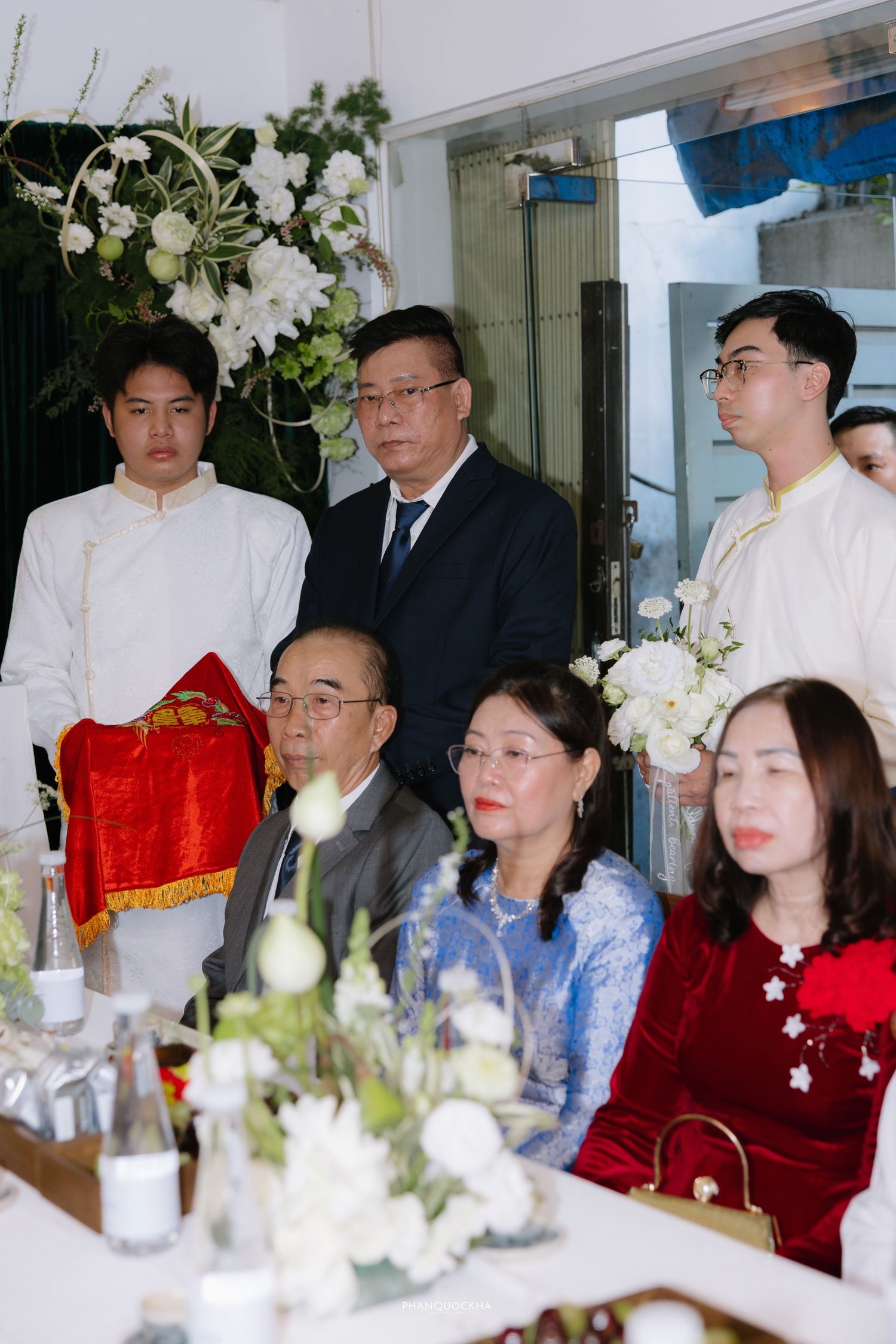 Wedding party members in ao dai standing behind seated family guests at traditional Vietnamese engagement ceremony.
