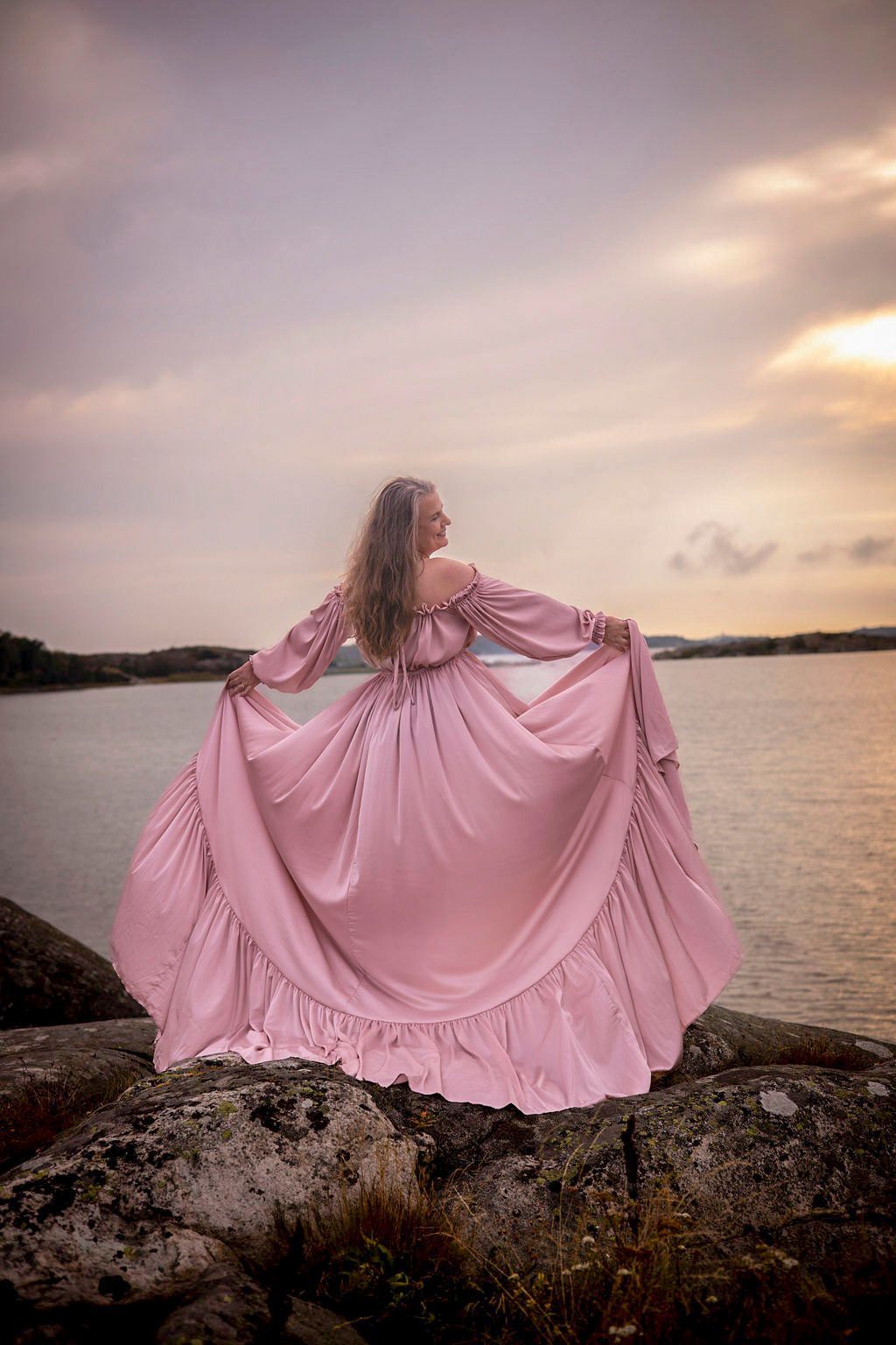bild tagen på en klippa med en kvinna som står ut mot havet iklädd en rosa klänning som hon håller ut åt sidorna.
