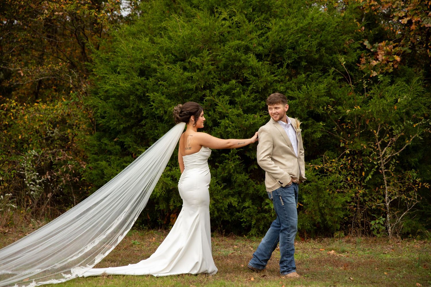 A person in a white wedding gown holds the hand of a small boy in jeans and suspenders in a forest setting.