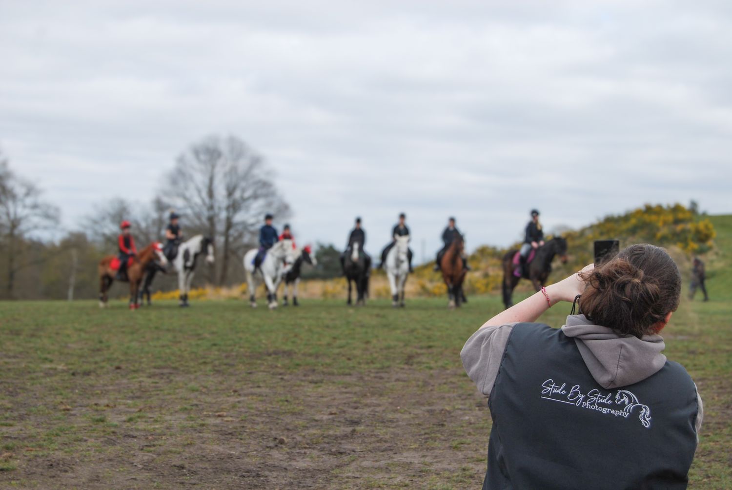 A photographer captures a group of horse riders gathered in an open field on a cloudy day.