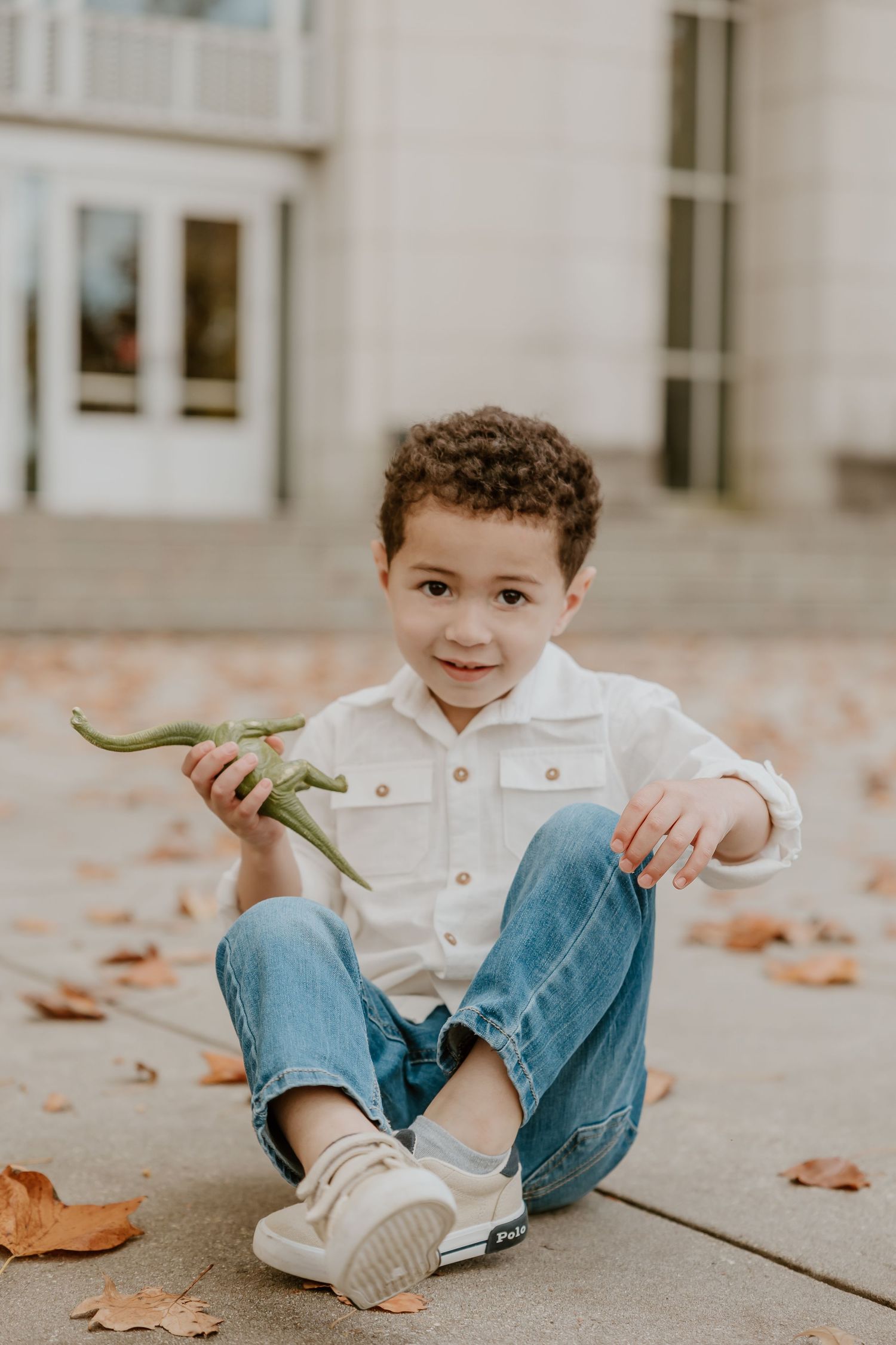 A young child sits on concrete steps wearing jeans and a white shirt while holding autumn leaves in a warm toned photo.