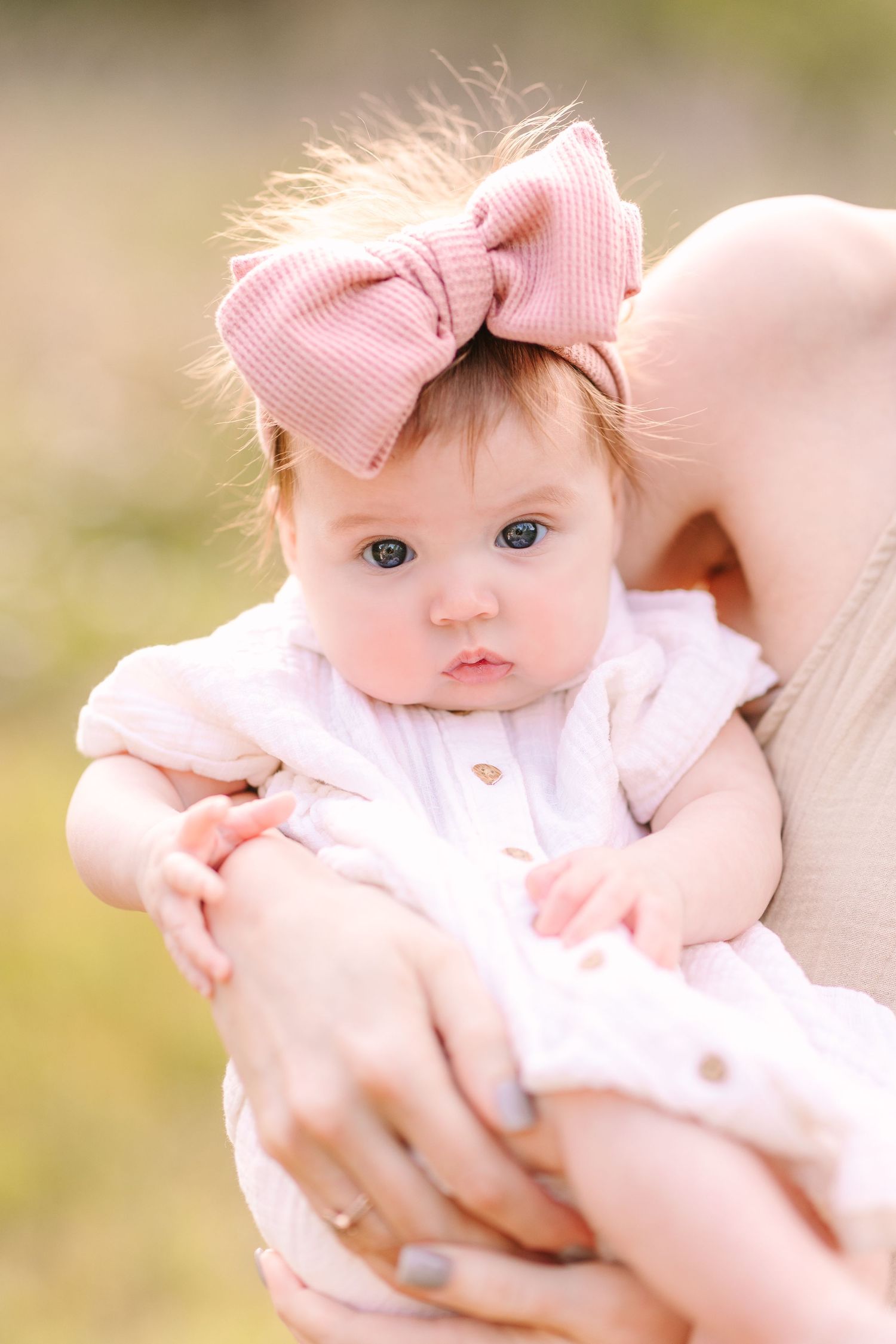 Sweet baby wearing pink bow headband lies against white blanket in outdoor portrait session.