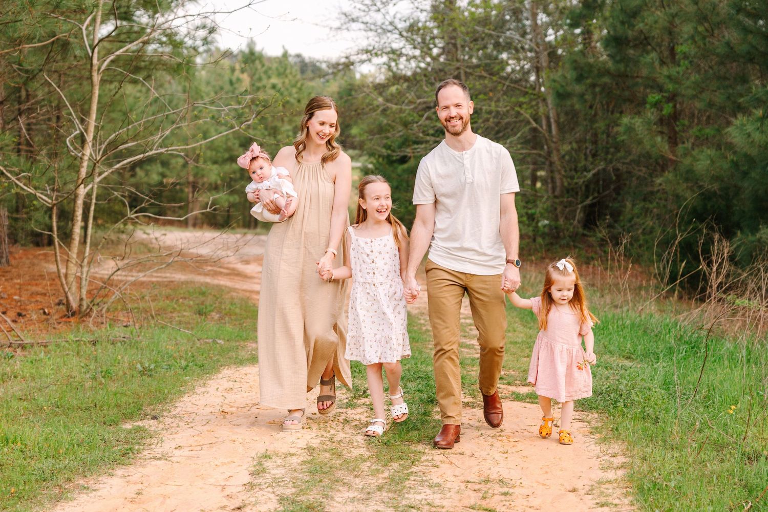 People in light colored clothing walk along a dirt path through a lush green countryside setting.