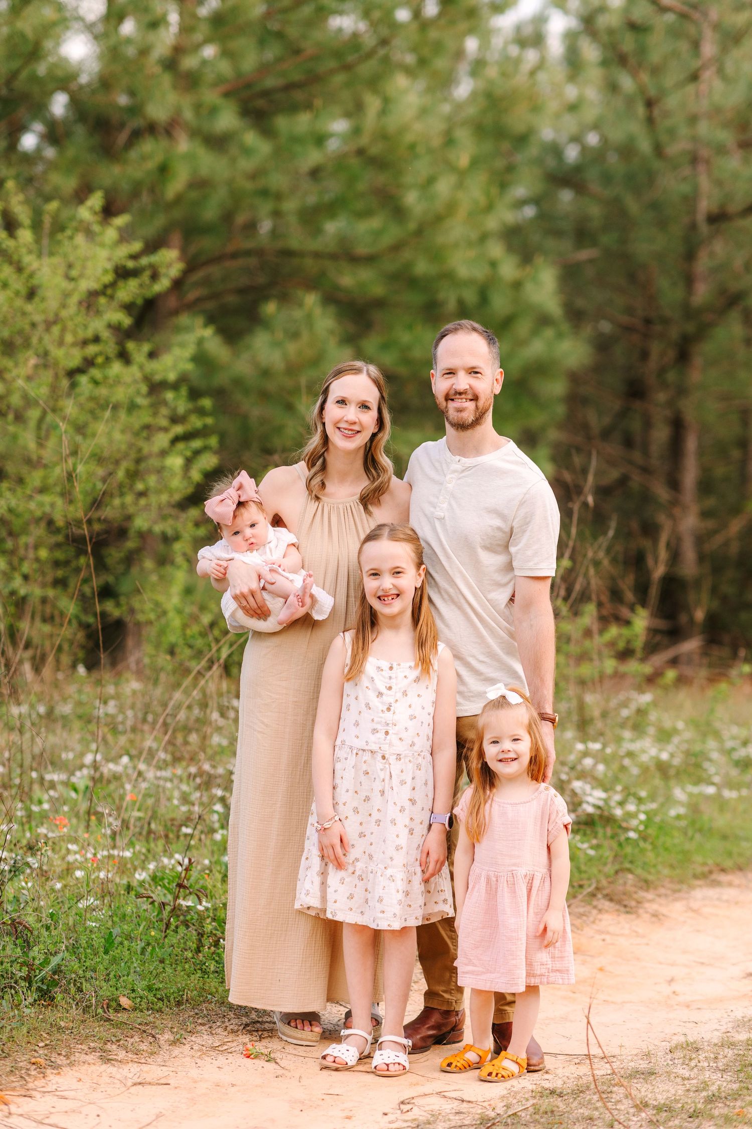 A family dressed in soft pink and white tones poses together on a dirt path surrounded by greenery and wildflowers.