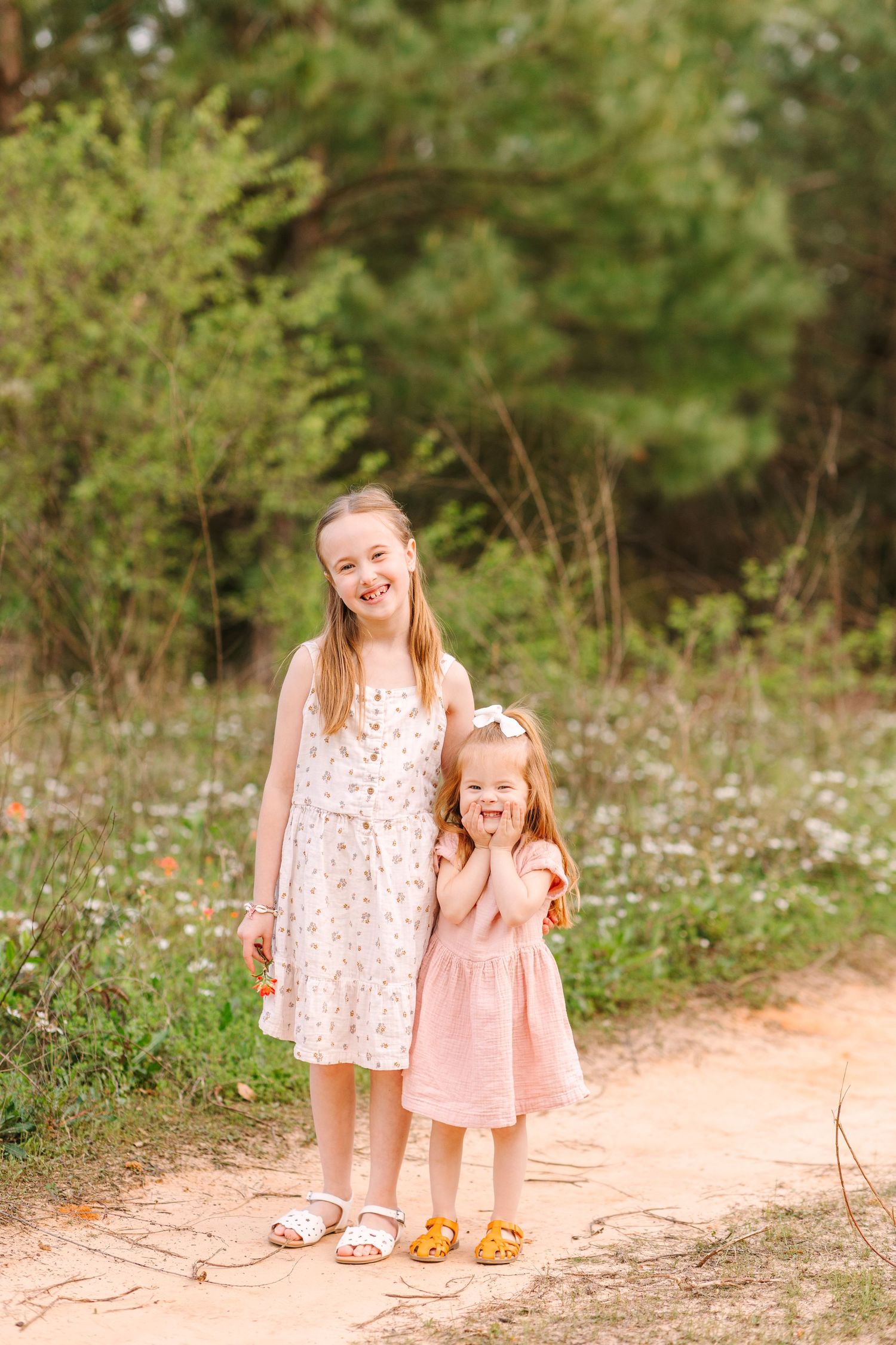 Two figures in pink dresses walking together on a dirt path surrounded by green foliage on a sunny day.