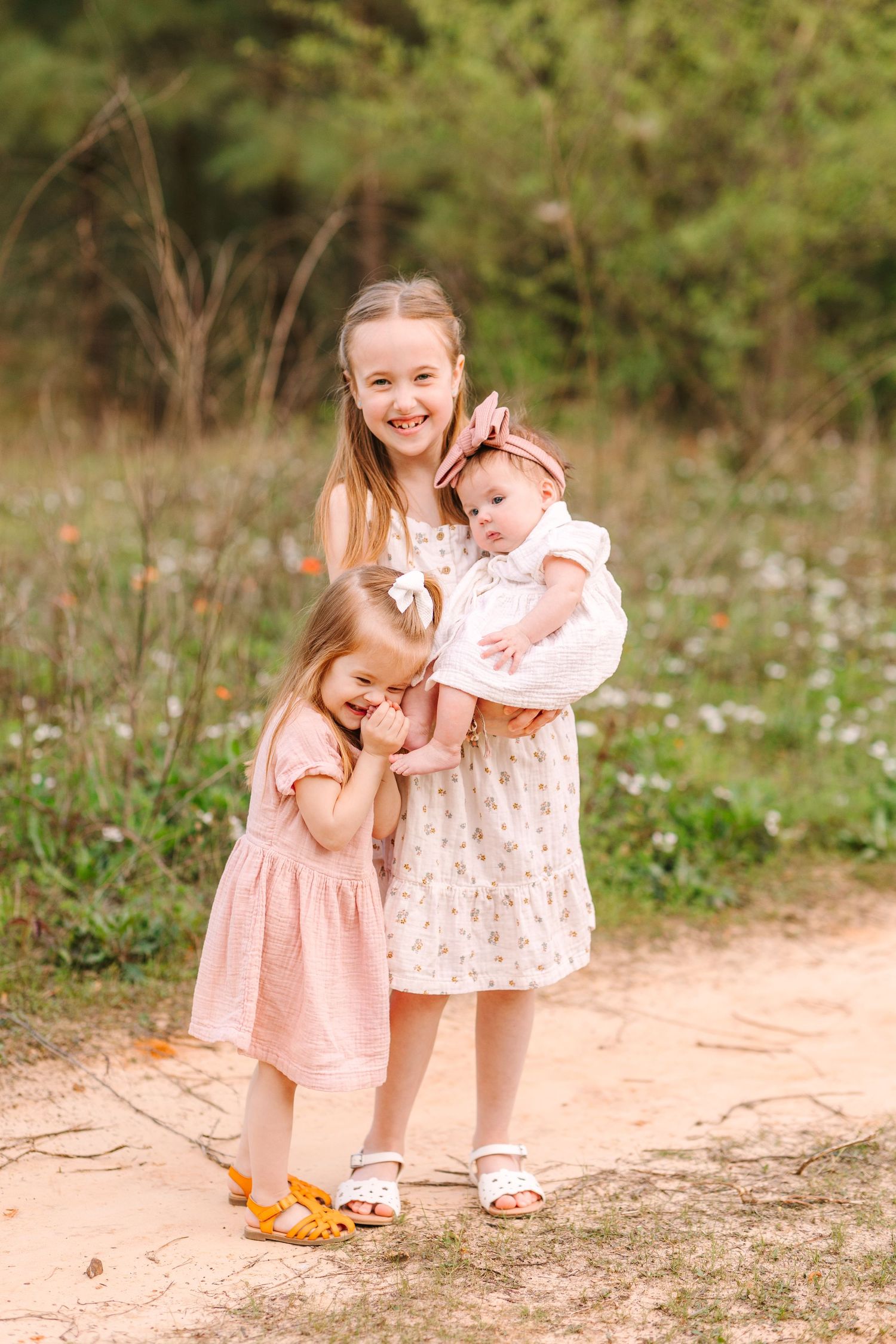 Two children in pink dresses embrace while standing on a dirt path surrounded by nature.